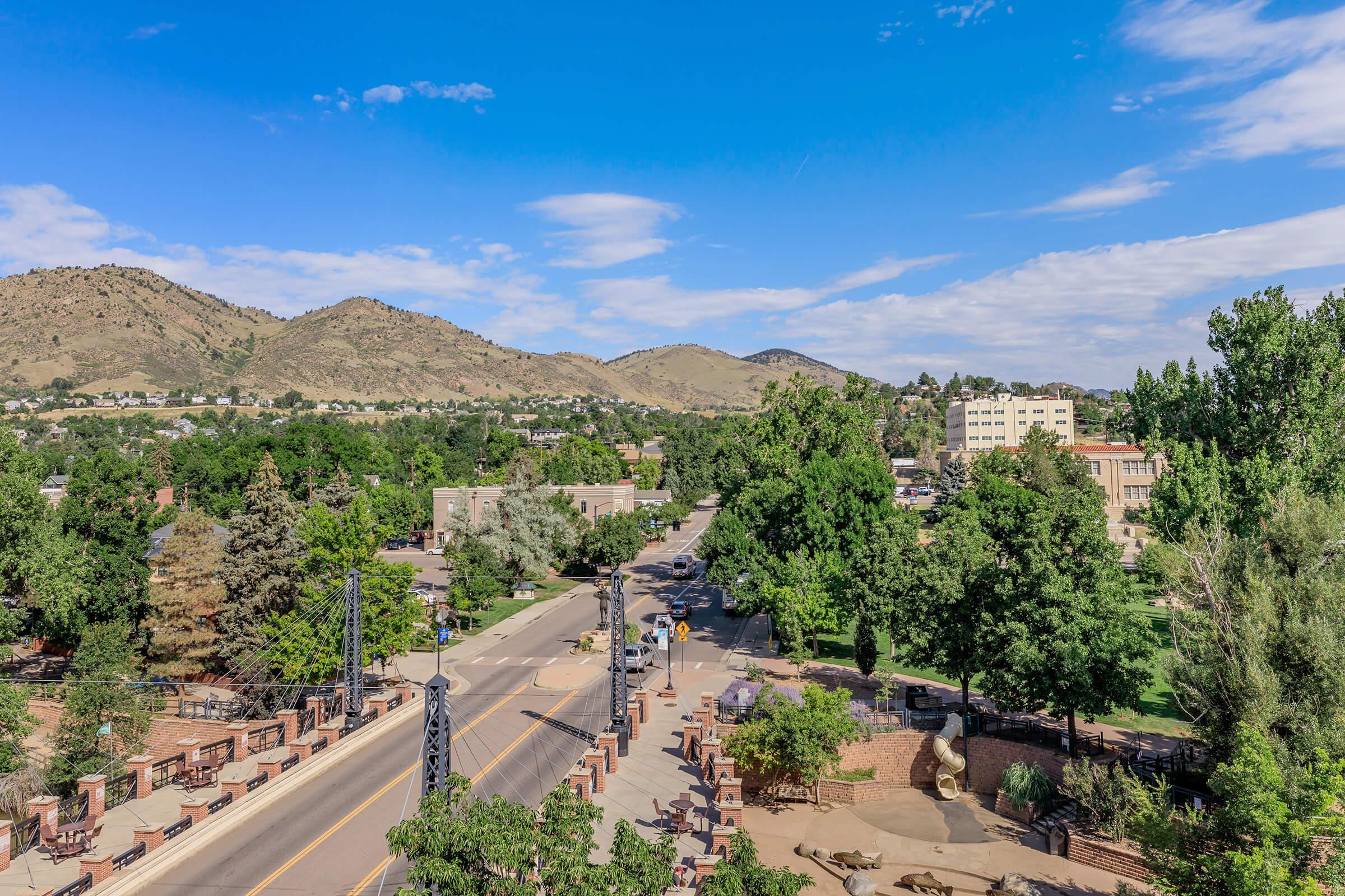 A wide aerial view of a scenic landscape featuring a clear blue sky and fluffy white clouds. In the foreground, a tree-lined street runs through a lush green area, with buildings and hills in the background. The scene is vibrant, showcasing nature and urban elements coexisting peacefully.