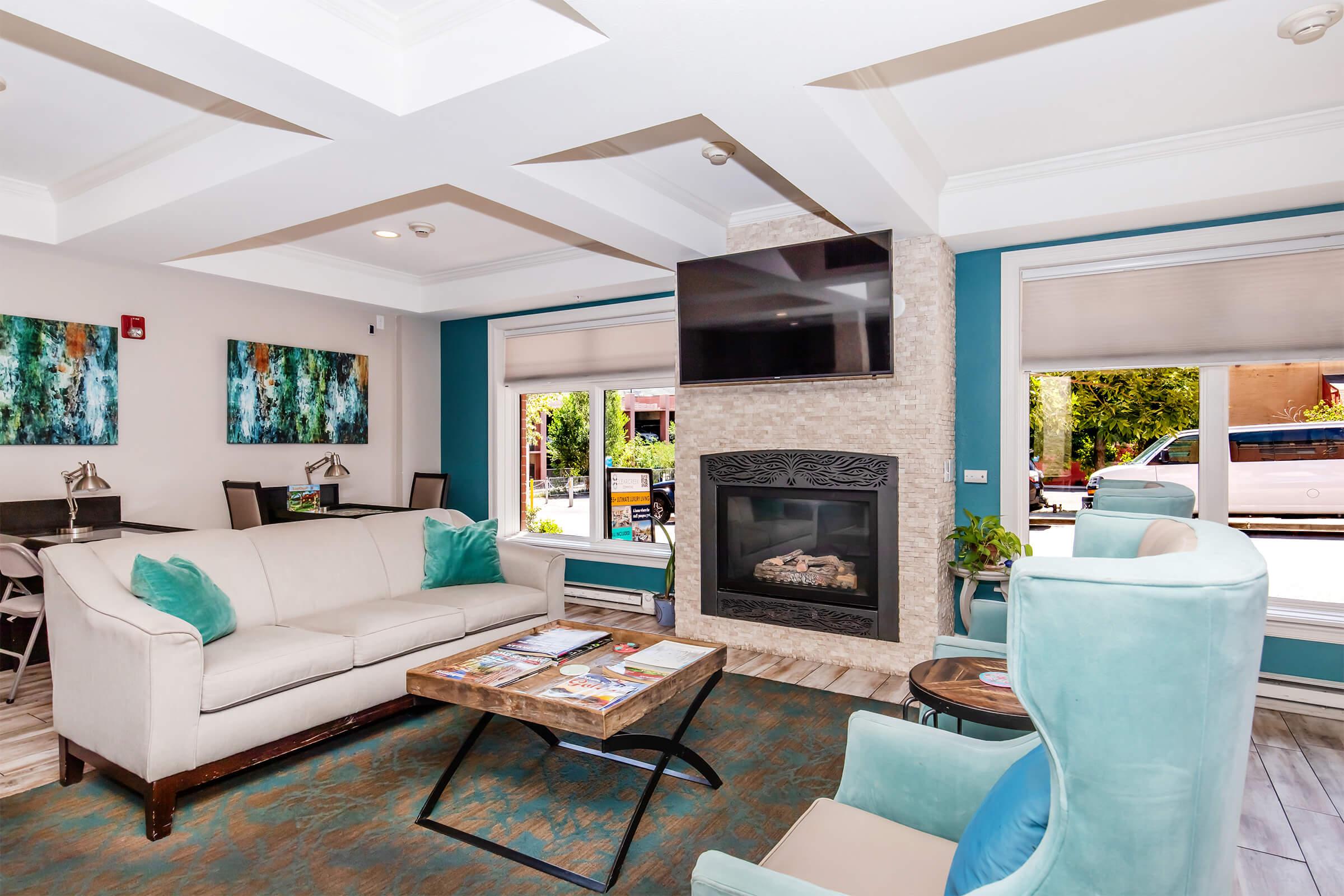 A cozy lounge area featuring a light-colored sofa, turquoise armchairs, and a stone fireplace. The walls are adorned with abstract art, and large windows allow natural light to flood the space. A flat-screen TV is mounted above the fireplace, creating a welcoming and stylish atmosphere for relaxation.