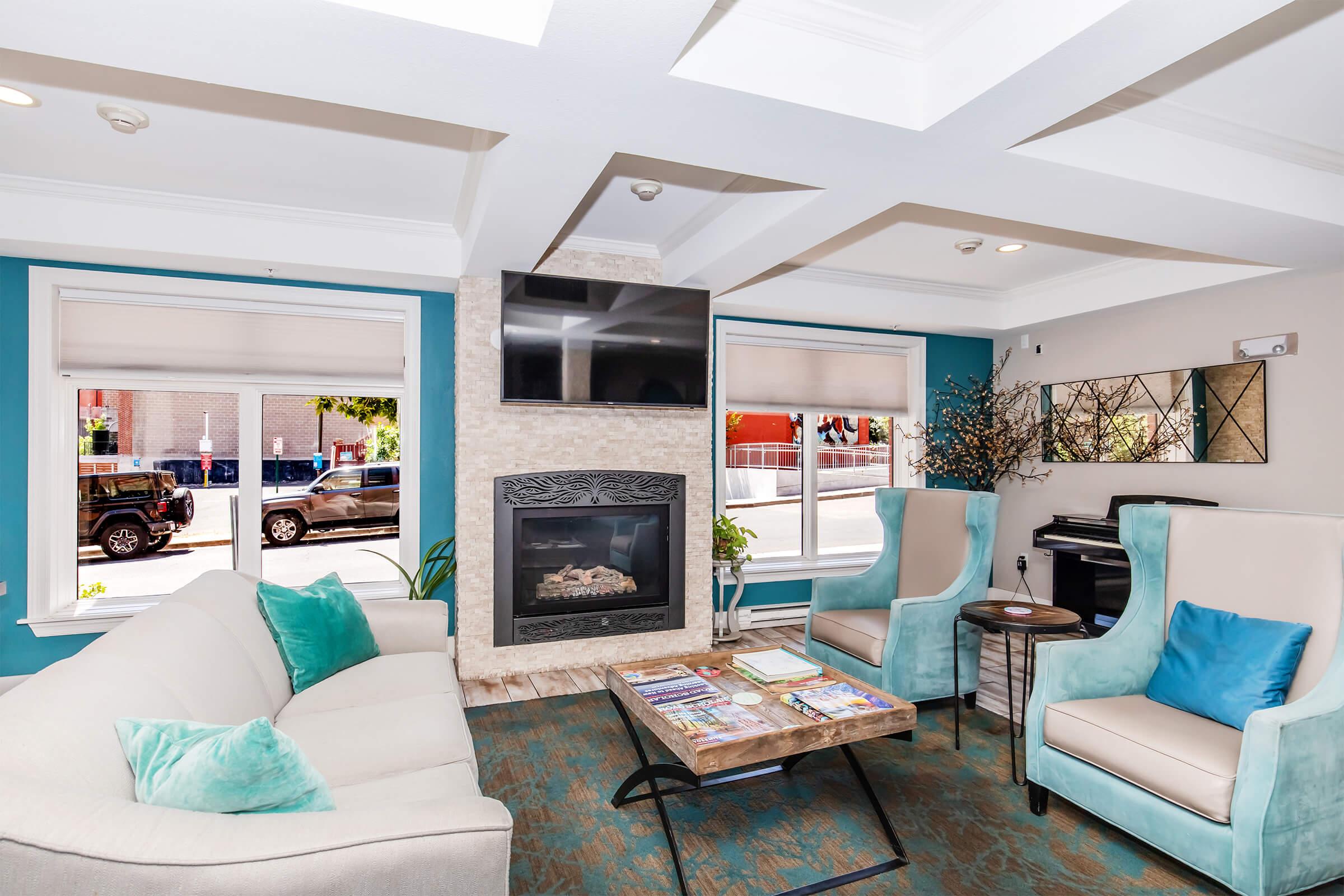 Stylish living room featuring a light-colored sofa with turquoise pillows, two modern armchairs, a decorative fireplace with a TV above it, and a wooden coffee table with magazines. Large windows provide natural light, and the walls are painted in blue and neutral tones, creating a welcoming atmosphere.