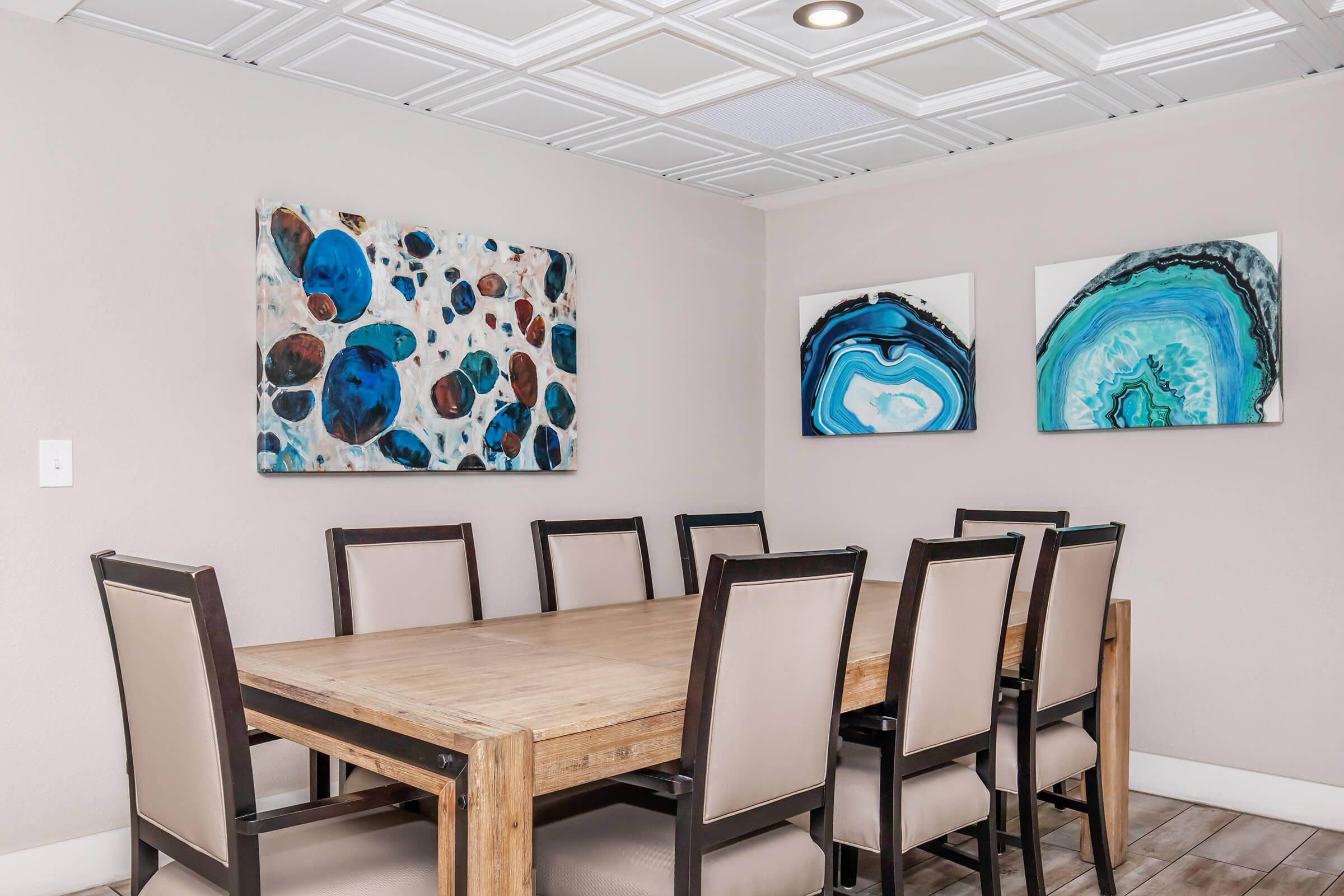 A modern dining room featuring a large wooden table surrounded by eight upholstered chairs. On the walls, there are two abstract paintings in shades of blue and green, alongside a colorful abstract piece with various circular shapes. The ceiling has a textured design, adding to the contemporary feel of the space.