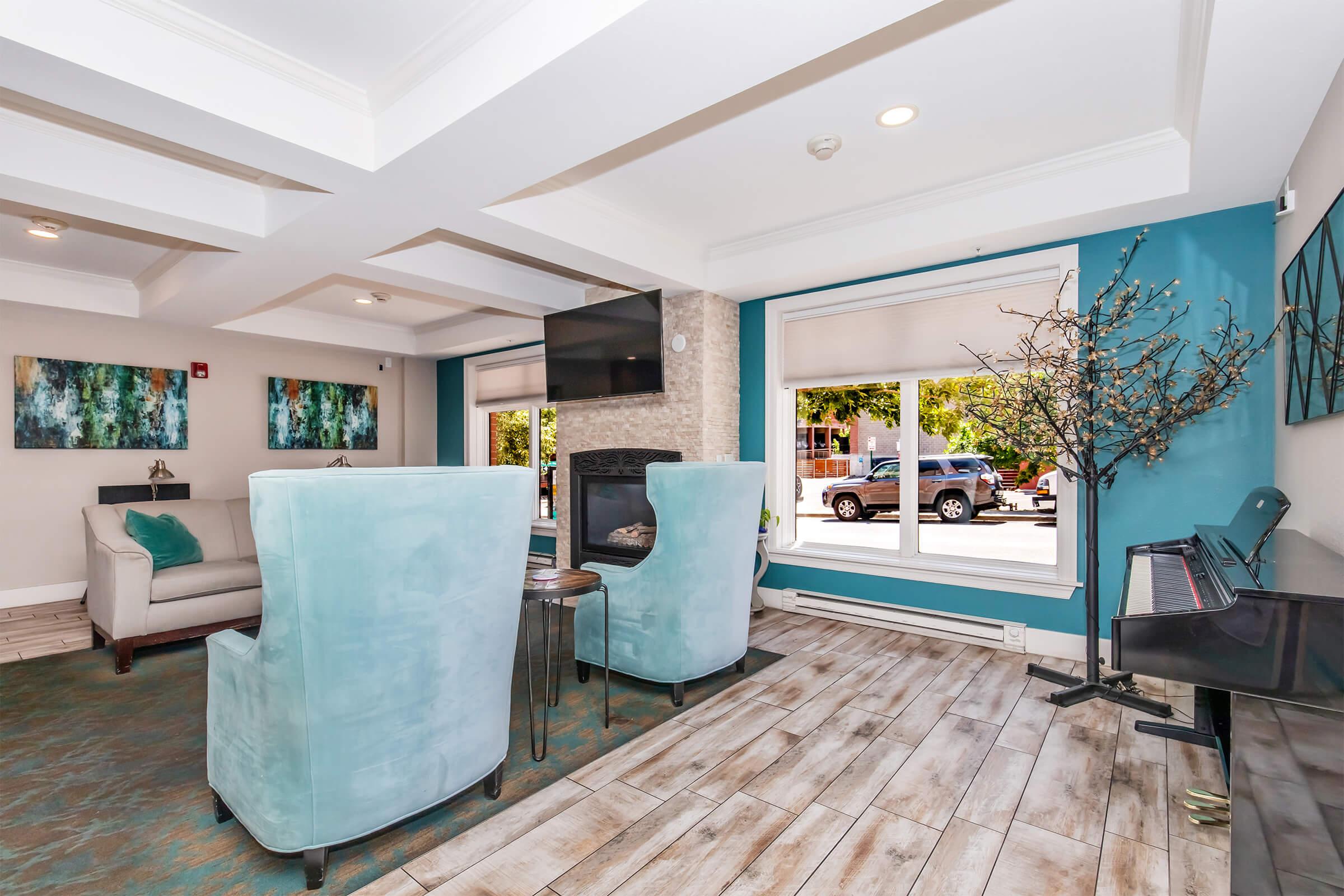 A cozy lounge area featuring light blue armchairs, a small round table, and a modern fireplace. Large windows let in natural light, overlooking outdoor scenery. A decorative branch adds a touch of nature, while a piano sits against the wall, enhancing the inviting atmosphere of the space.