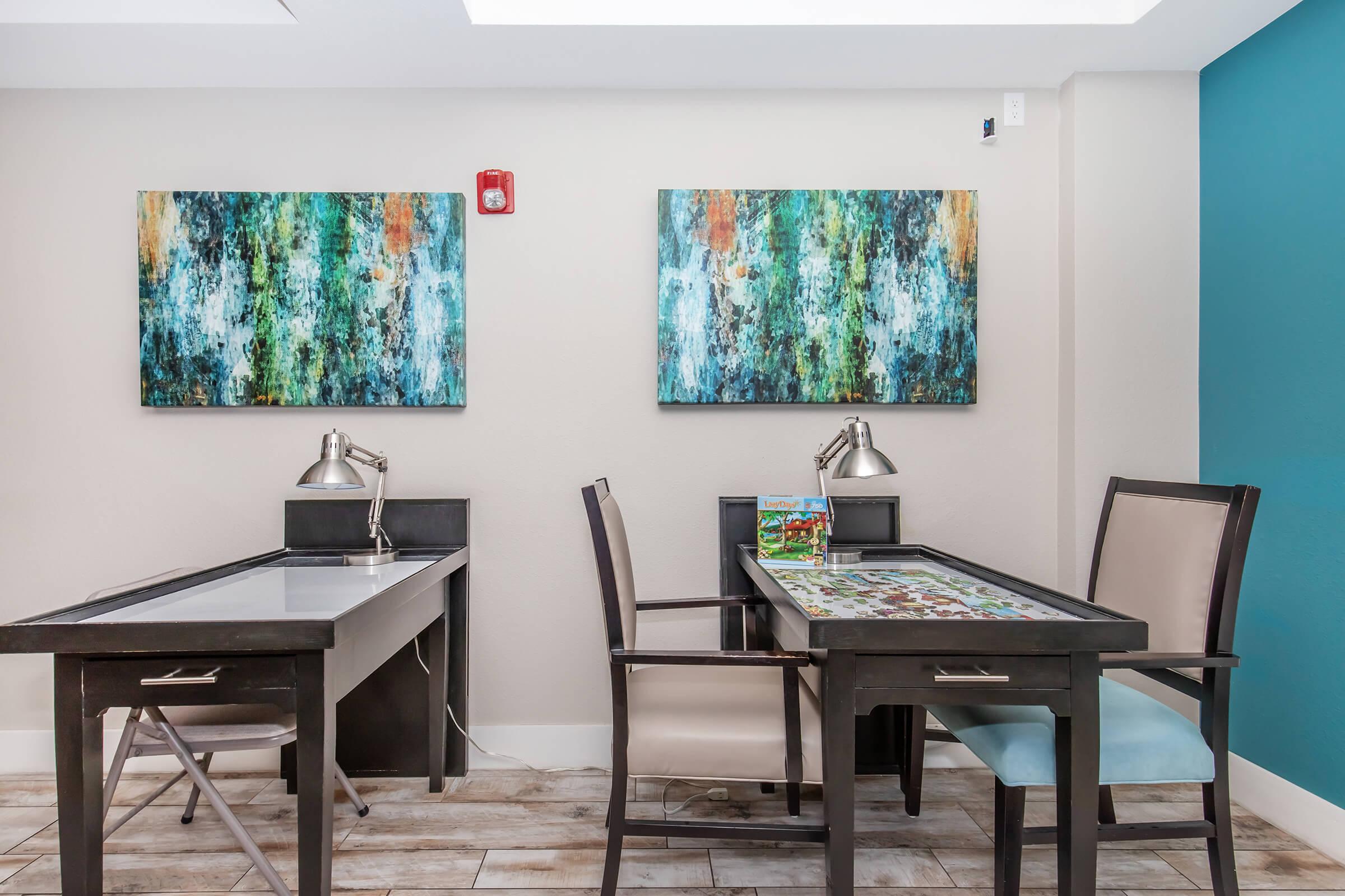 Two study desks with lamps in a well-lit room featuring colorful abstract art on the walls. One desk has a puzzle partially completed, while the other is empty. The room is decorated in calming colors, combining teal and neutral tones, creating a welcoming and functional workspace.