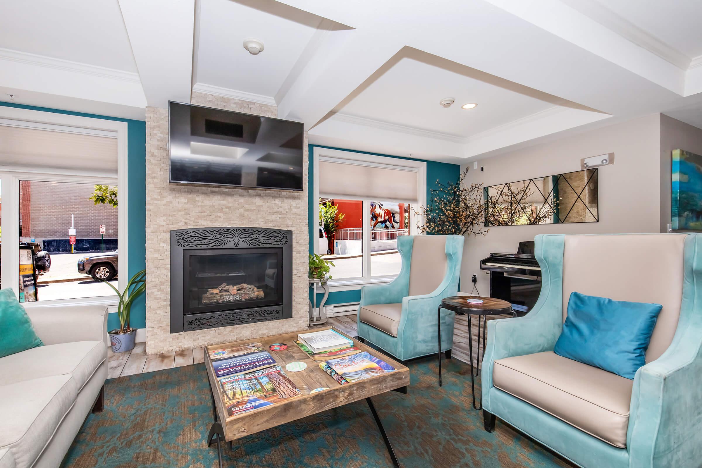 A modern lounge area featuring two light blue upholstered chairs and a wooden coffee table with magazines. A flat-screen TV is mounted above a decorative stone fireplace. Large windows provide natural light, and there's a plant in the corner, creating a cozy and inviting atmosphere.