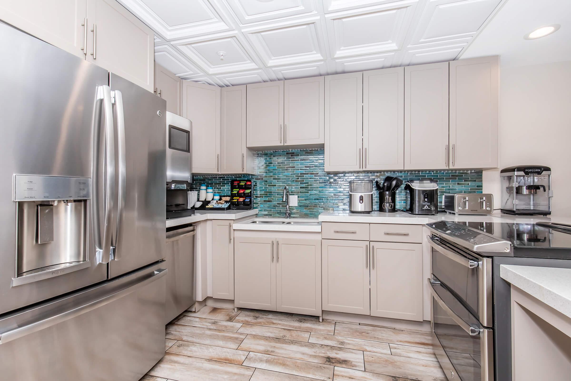 Modern kitchen featuring stainless steel appliances, including a refrigerator and oven. Light-colored cabinets are paired with a blue mosaic backsplash. The countertop is white, and various kitchen appliances, like a coffee maker and a toaster, are visible. The flooring consists of wood-like tiles.