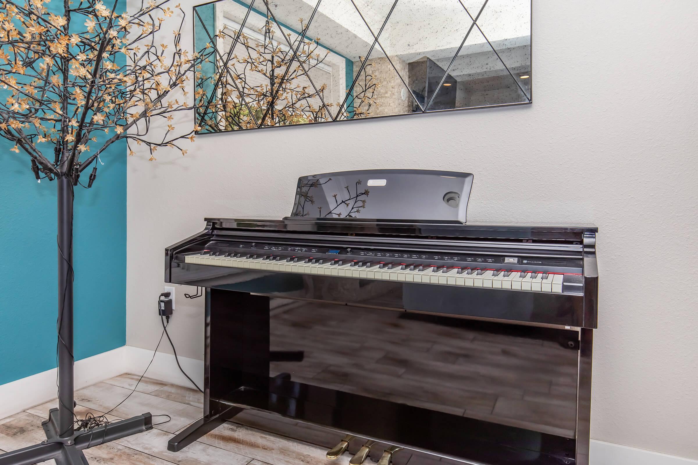 A black digital piano with a closed lid, positioned against a wall. Above it is a large mirror reflecting the space. Next to the piano, there is a decorative tree with LED lights, adding a warm ambiance to the room. The walls are painted in a soft teal color, and the flooring is a light wood.