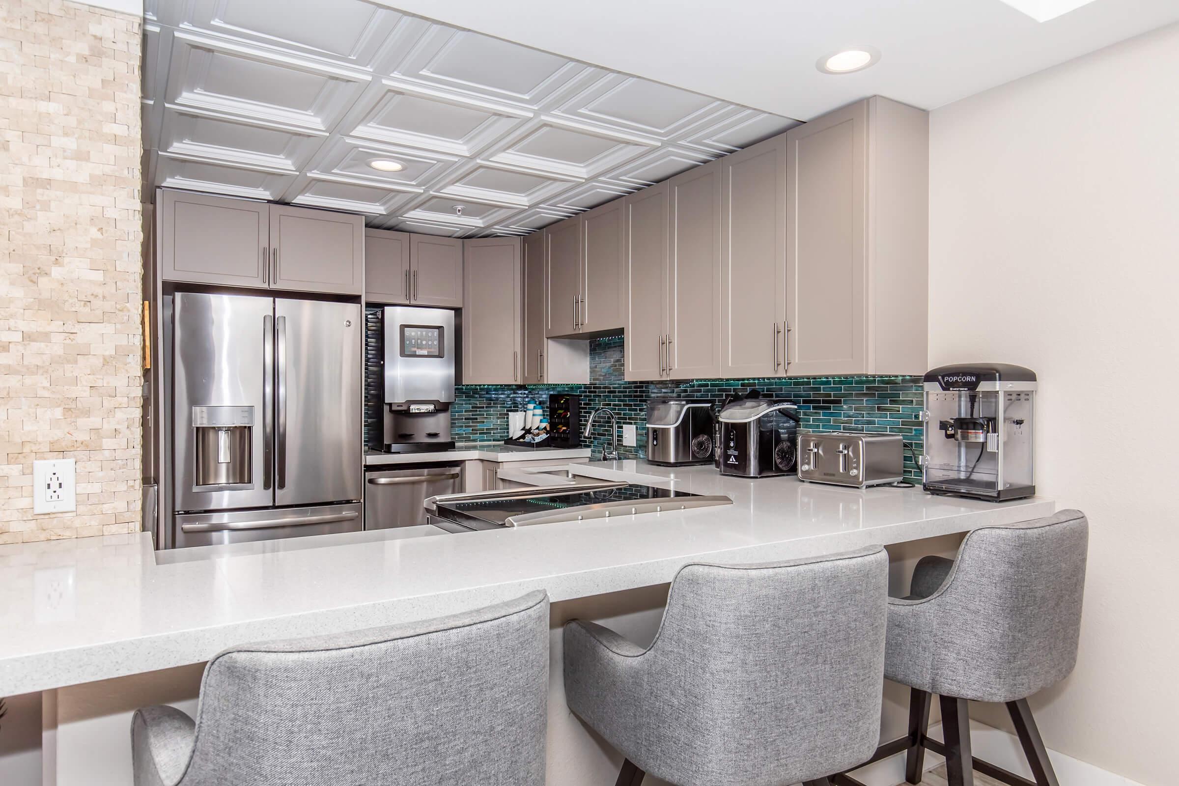 Modern kitchen with stainless steel appliances, including a refrigerator, oven, and coffee machine. The cabinets are a light gray color, and there's a blue mosaic tile backsplash. A sleek white countertop is paired with gray bar stools, creating a stylish and functional space for cooking and dining.