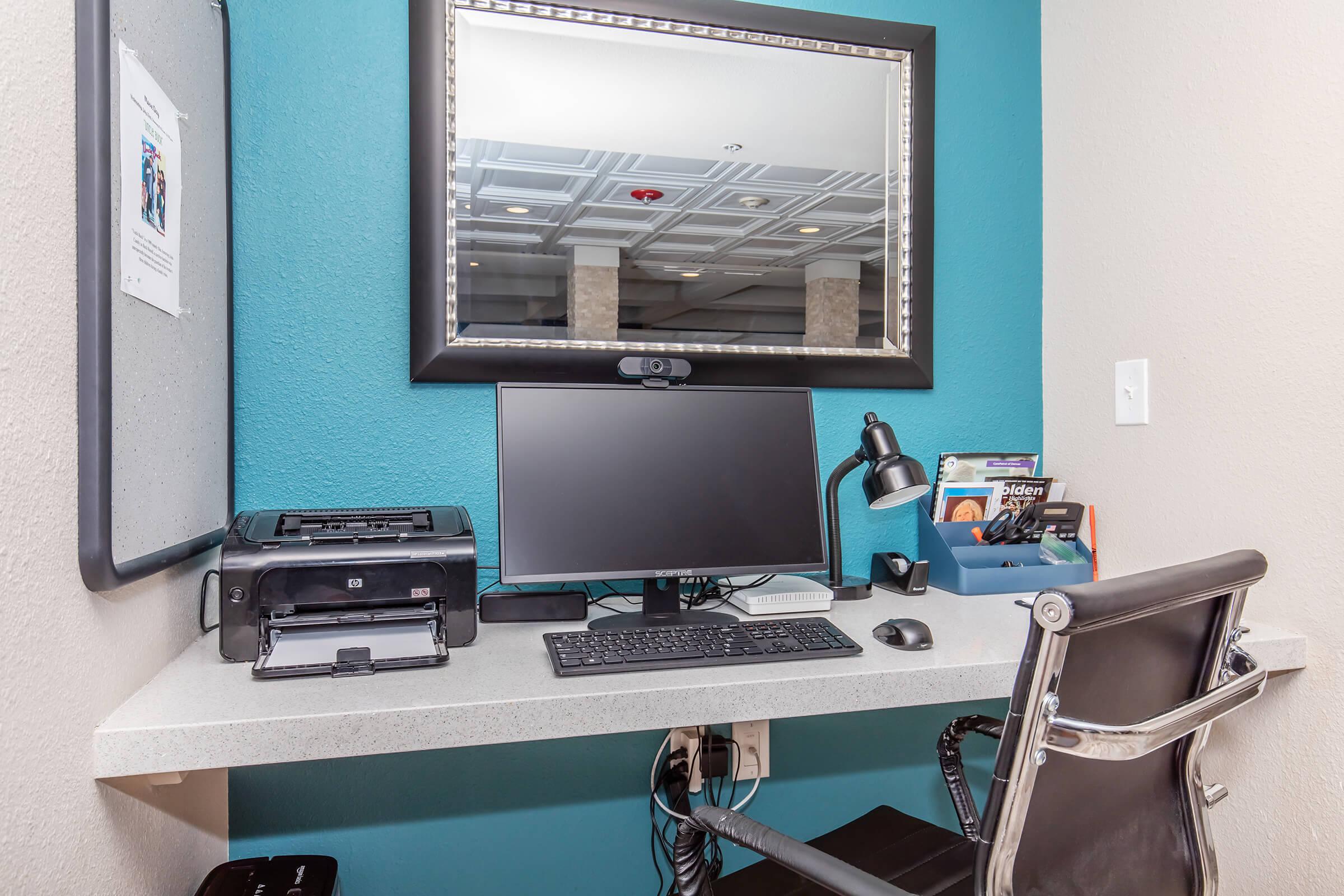 A modern workspace featuring a computer monitor, keyboard, and a desk lamp on a light-colored desk. There's a printer on the left and a blue organizer holding documents. A large mirror is mounted on the wall above the desk, with a teal accent wall in the background. A sleek chair is positioned in front of the desk.