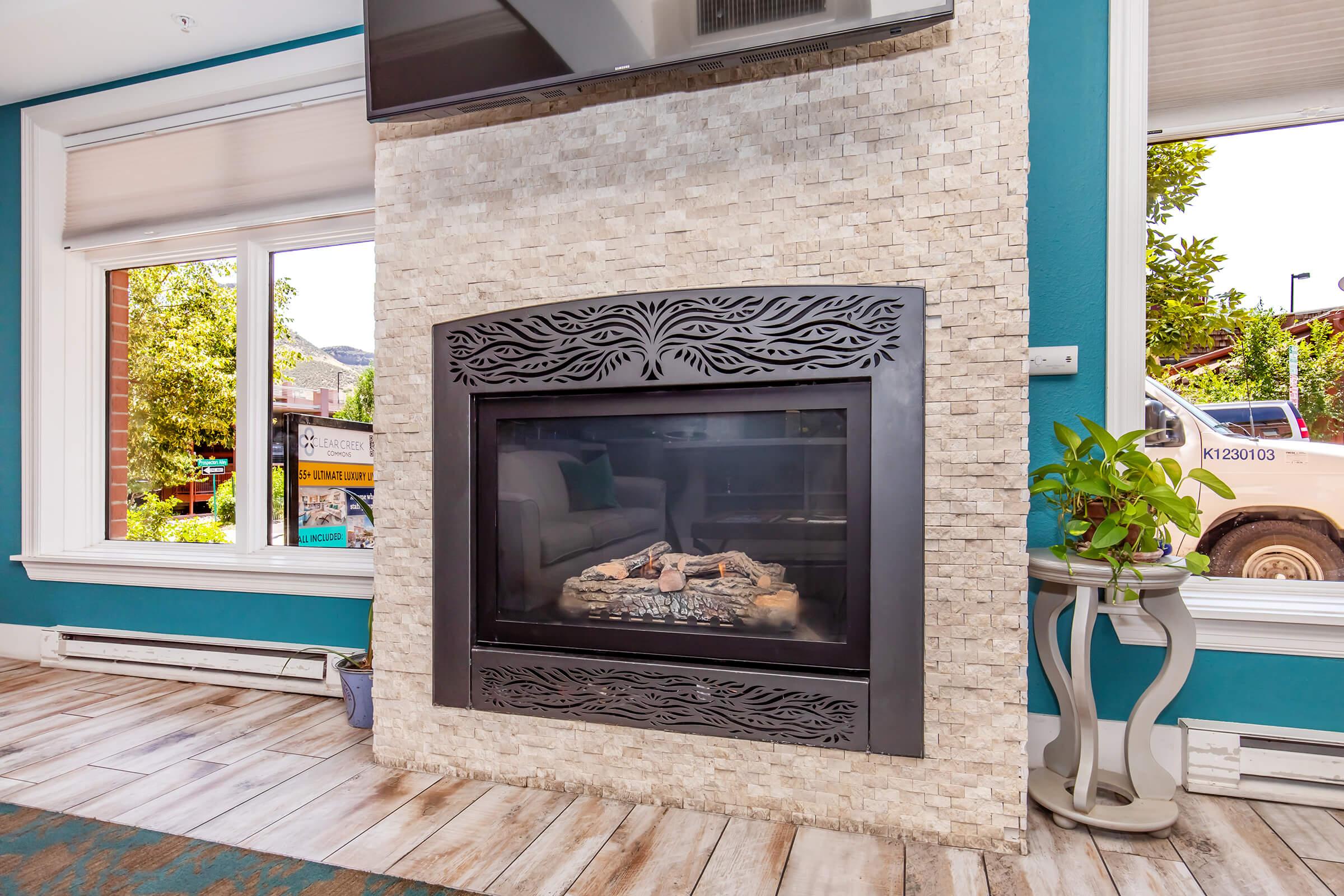 A modern fireplace with an ornate black metallic frame is set within a textured stone wall. The surrounding room features turquoise walls and large windows that let in natural light, showcasing greenery outside. A comfortable seating area is partially visible, creating a cozy atmosphere.