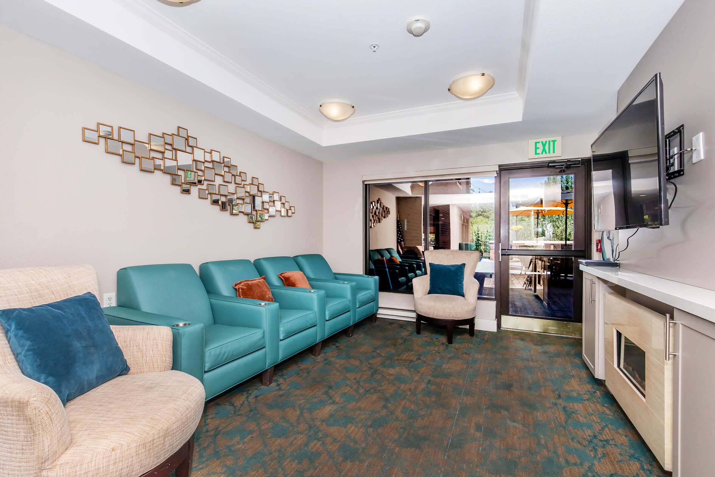 A modern lounge area featuring teal armchairs with orange and blue cushions, a beige armchair, a wall decorated with geometric mirrors, and a large flat-screen TV. A glass door leads to an outdoor space, while the floor has a textured carpet in muted tones. Bright lighting enhances the cozy atmosphere.