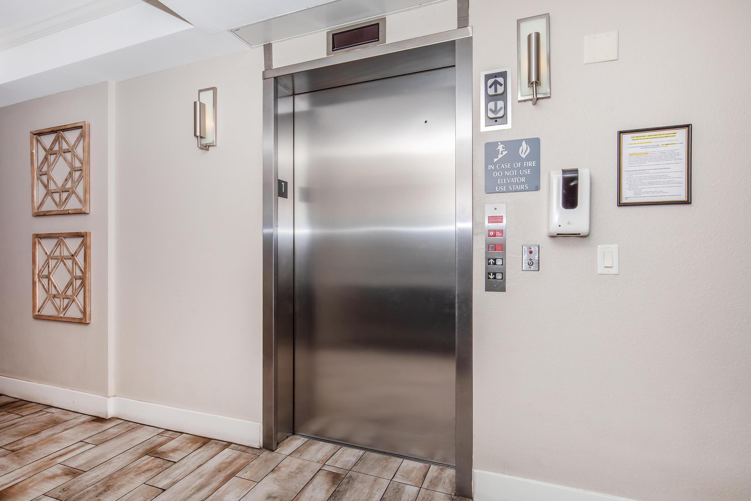 Elevator in a hallway with modern decor. The elevator doors are shiny and metallic, and there are several buttons and signs nearby, including instructions and emergency information. The walls are painted a neutral color, and there are decorative wall hangings framing the elevator area.