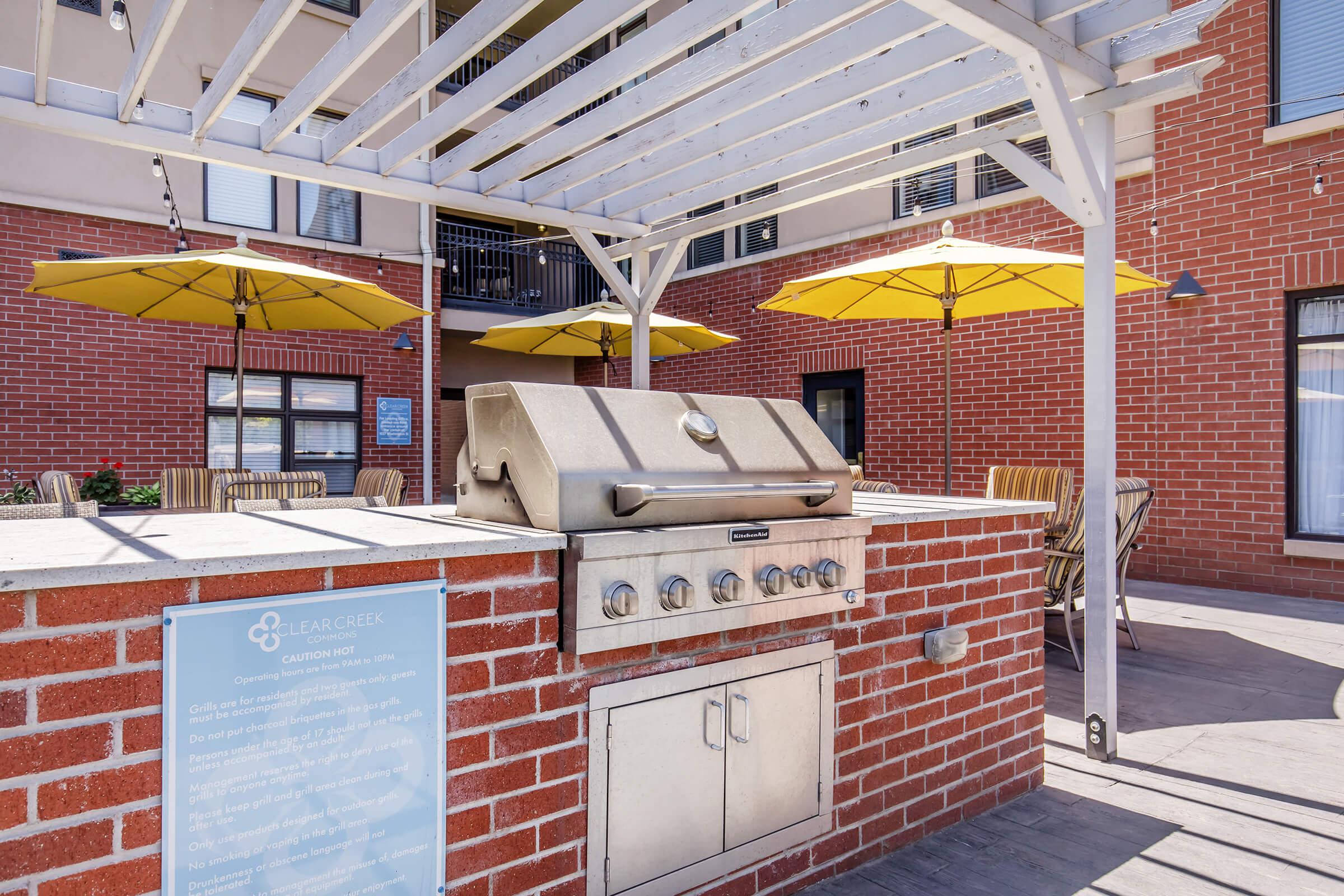 An outdoor grilling area featuring a stainless steel gas grill built into a brick structure. Yellow umbrellas provide shade over nearby tables and chairs, creating a welcoming space for gatherings. The setting is well-lit and designed for socializing in a residential environment.