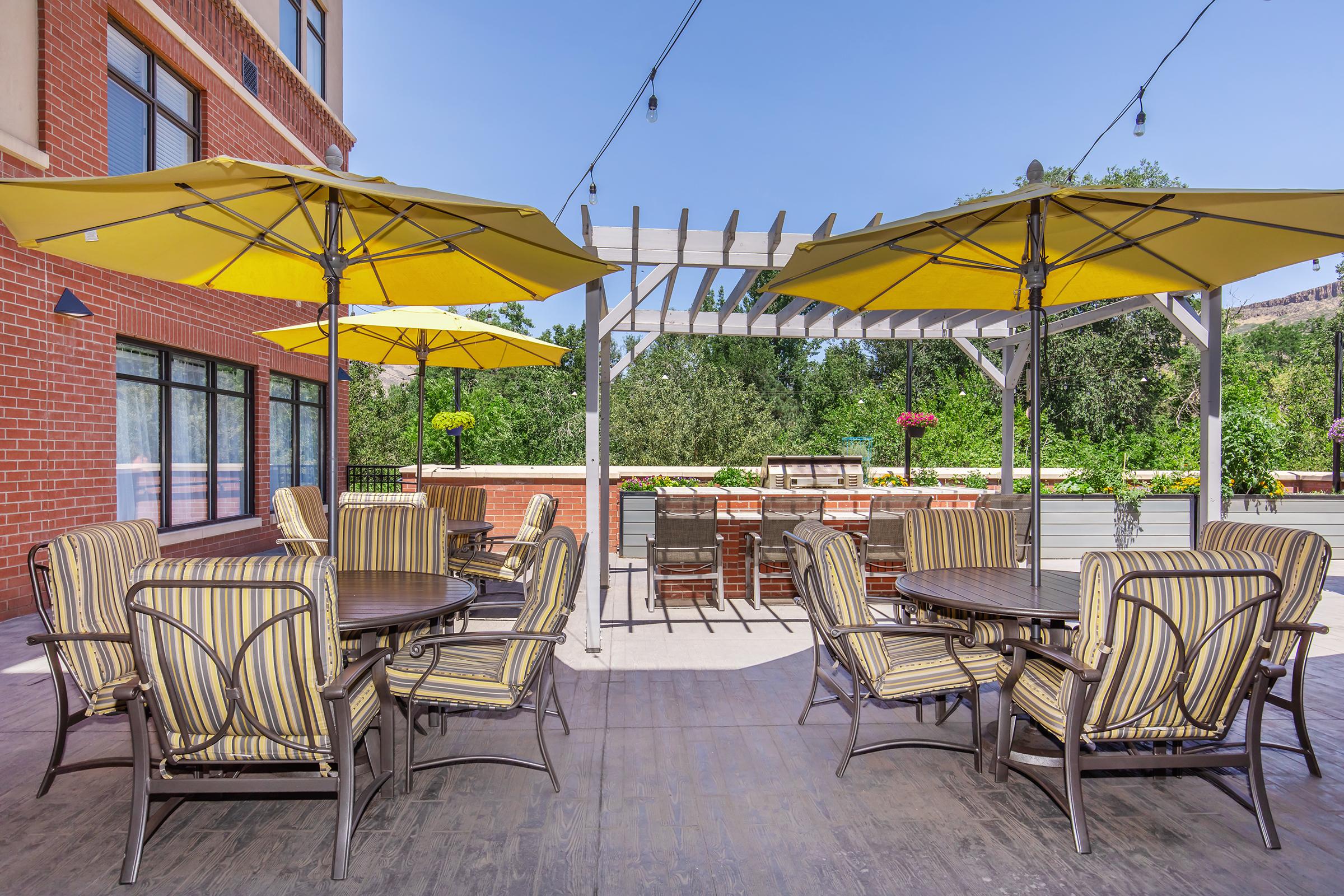A sunny outdoor patio featuring several round tables with striped yellow umbrellas. There are cushioned chairs around the tables, and a wooden pergola in the background. The area is surrounded by greenery and decorative plants, creating a lively and inviting atmosphere.