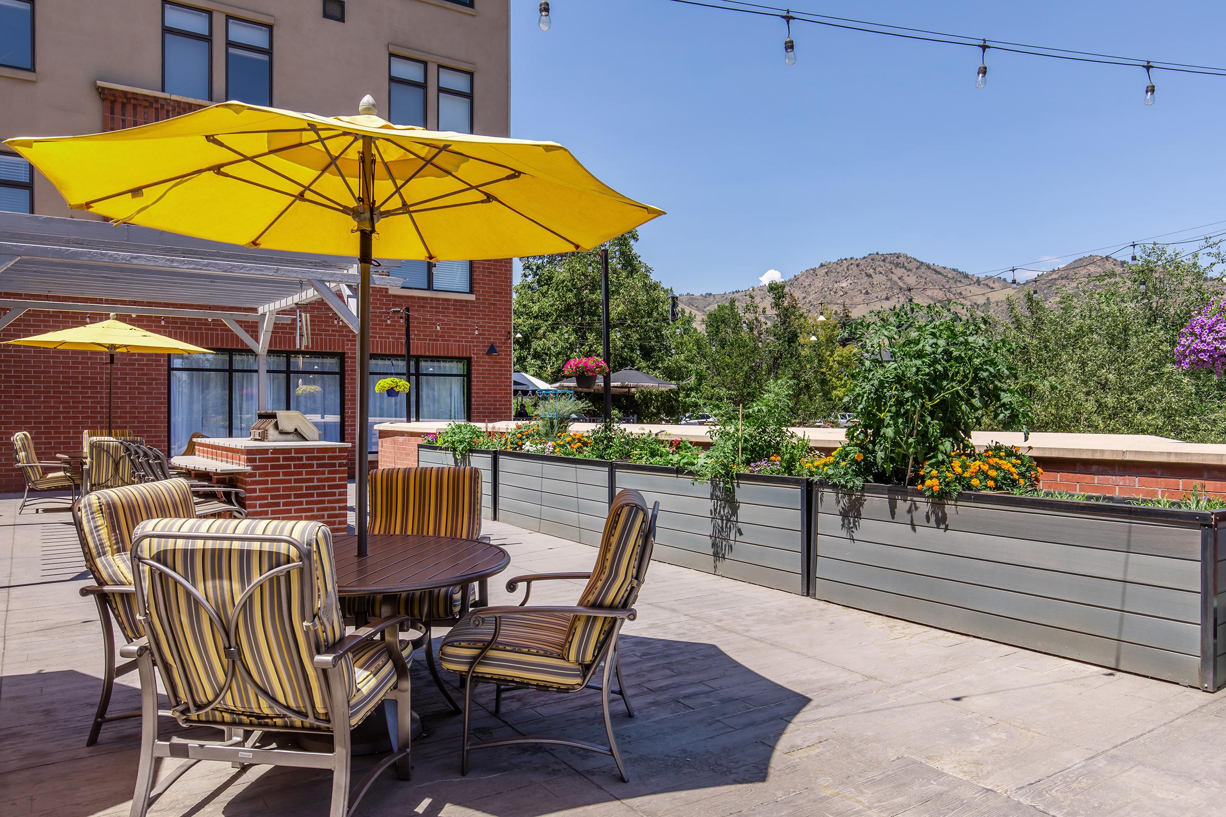 Outdoor dining area featuring a round wooden table surrounded by striped chairs under a bright yellow umbrella. The setting includes raised garden beds with blooming flowers and greenery, with mountains visible in the background and a clear blue sky overhead. String lights are hung above for ambiance.
