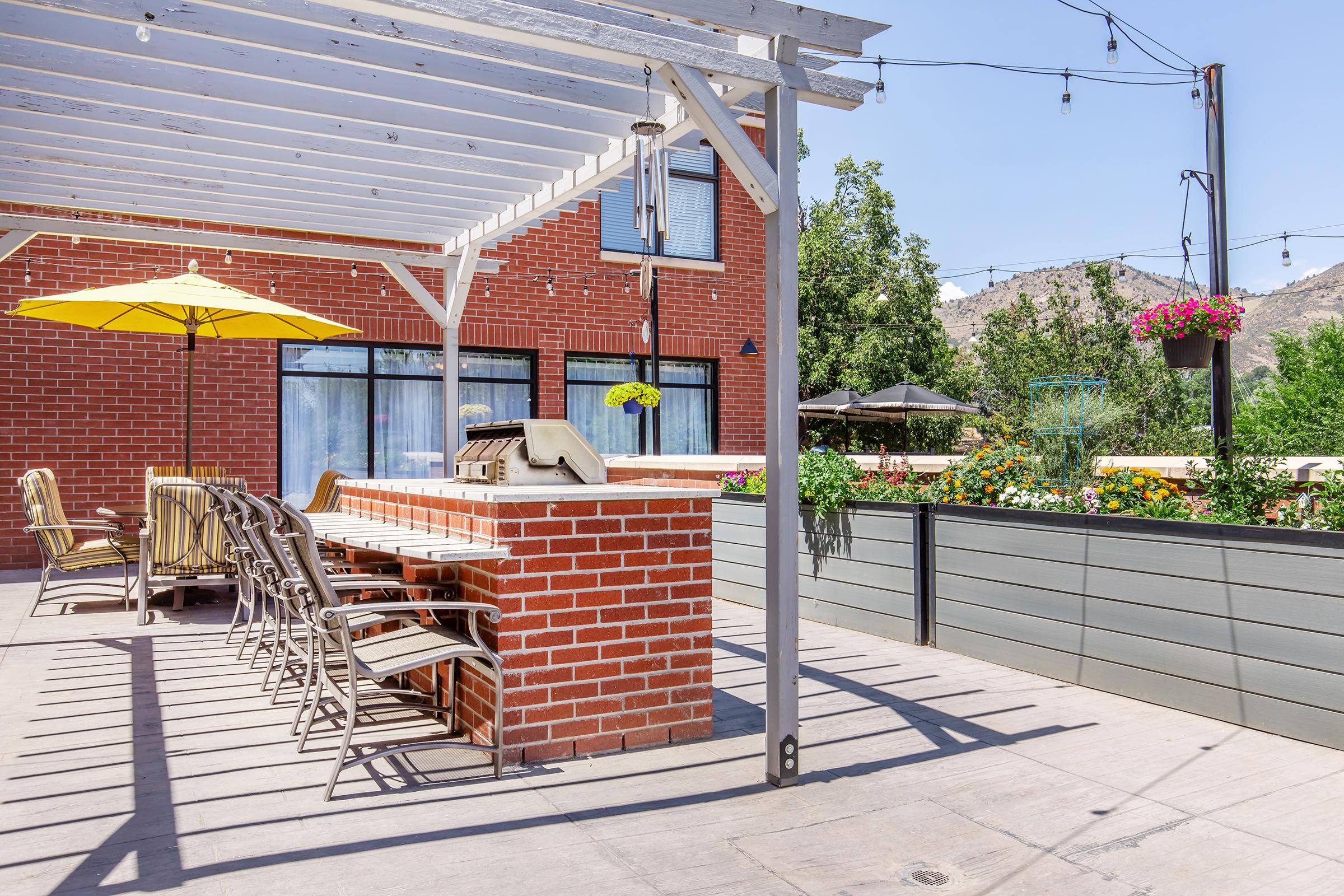 A patio area featuring a brick grill station, several outdoor chairs, and a yellow umbrella. The space is surrounded by lush greenery and colorful flowers, with string lights overhead. The background includes a simple brick building and mountains in the distance, creating a relaxed outdoor setting.