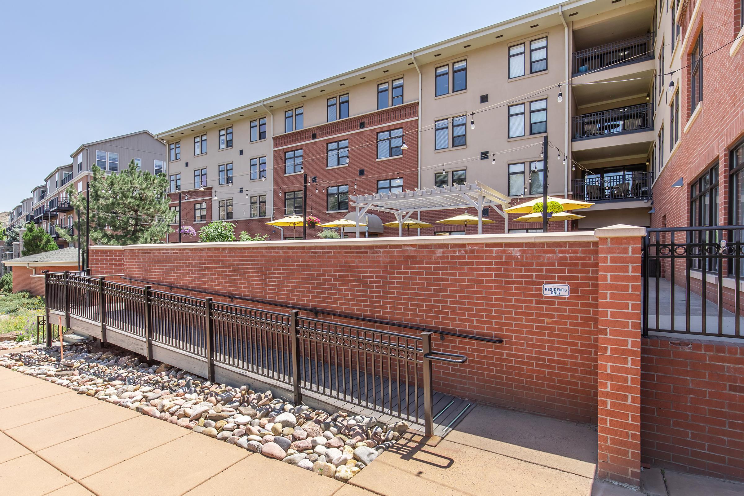 A well-maintained outdoor area of an apartment complex featuring a ramp leading to a spacious patio with yellow umbrellas. The building has a modern architectural design with a mix of brick and cream-colored siding, surrounded by landscaped greenery and natural stone accents on the pathway.