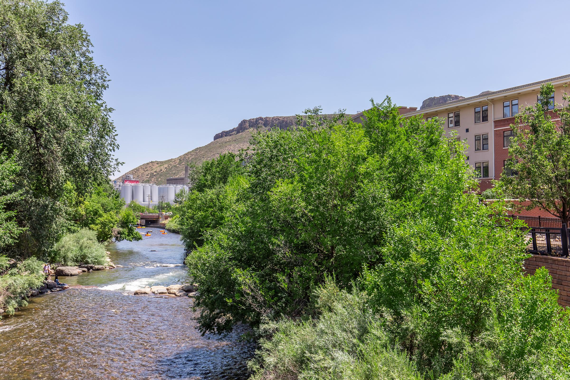 A serene river flows through a lush green landscape, flanked by dense trees and shrubs. In the background, a rocky mountain rises against a clear blue sky, while a building is partially visible on the right side, contributing to the picturesque natural setting.