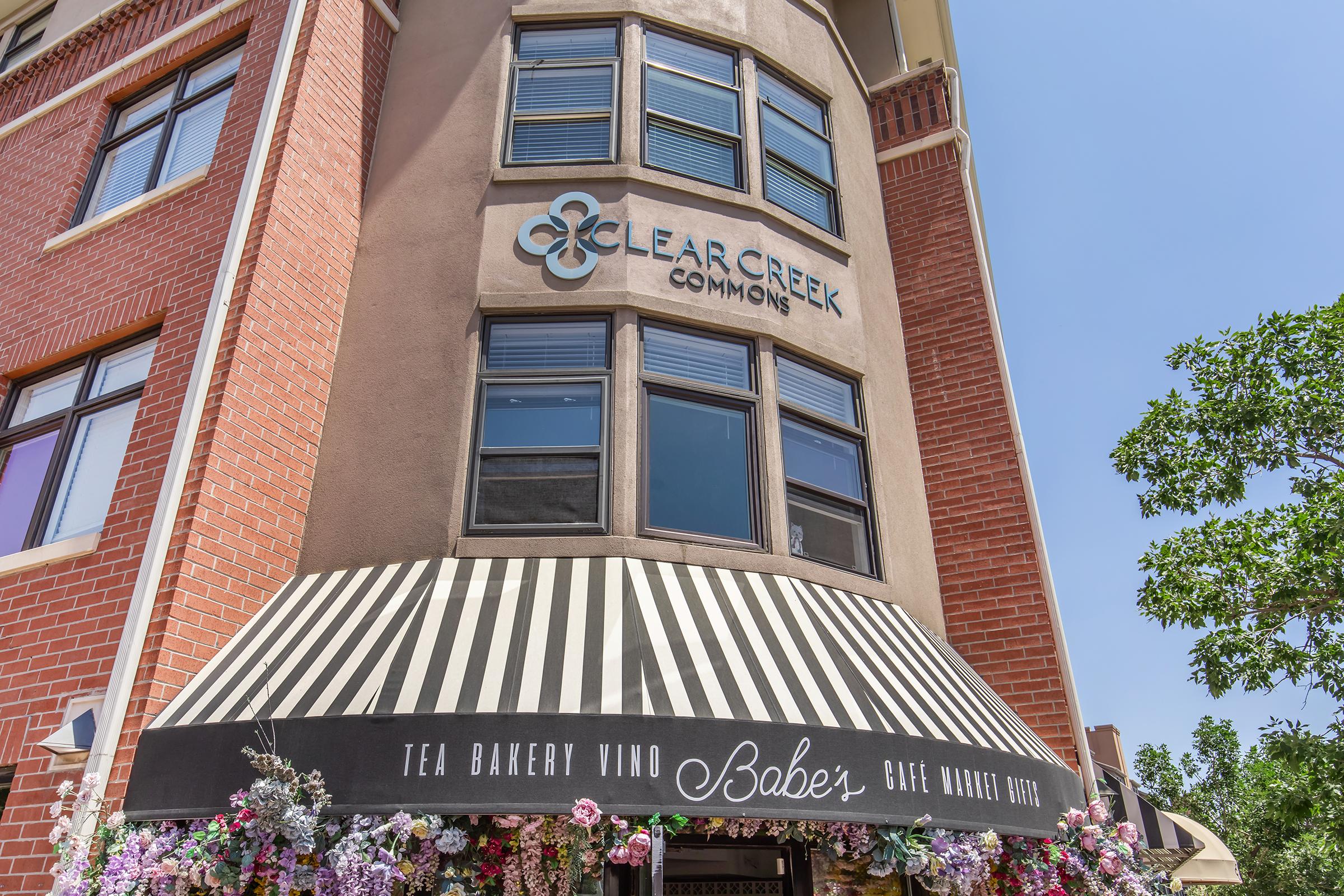 Building corner featuring the sign "Clear Creek Commons." Below, an awning displays "Babe's Tea Bakery Vino" with a floral arrangement. The architecture includes brick and contemporary windows, set against a blue sky.