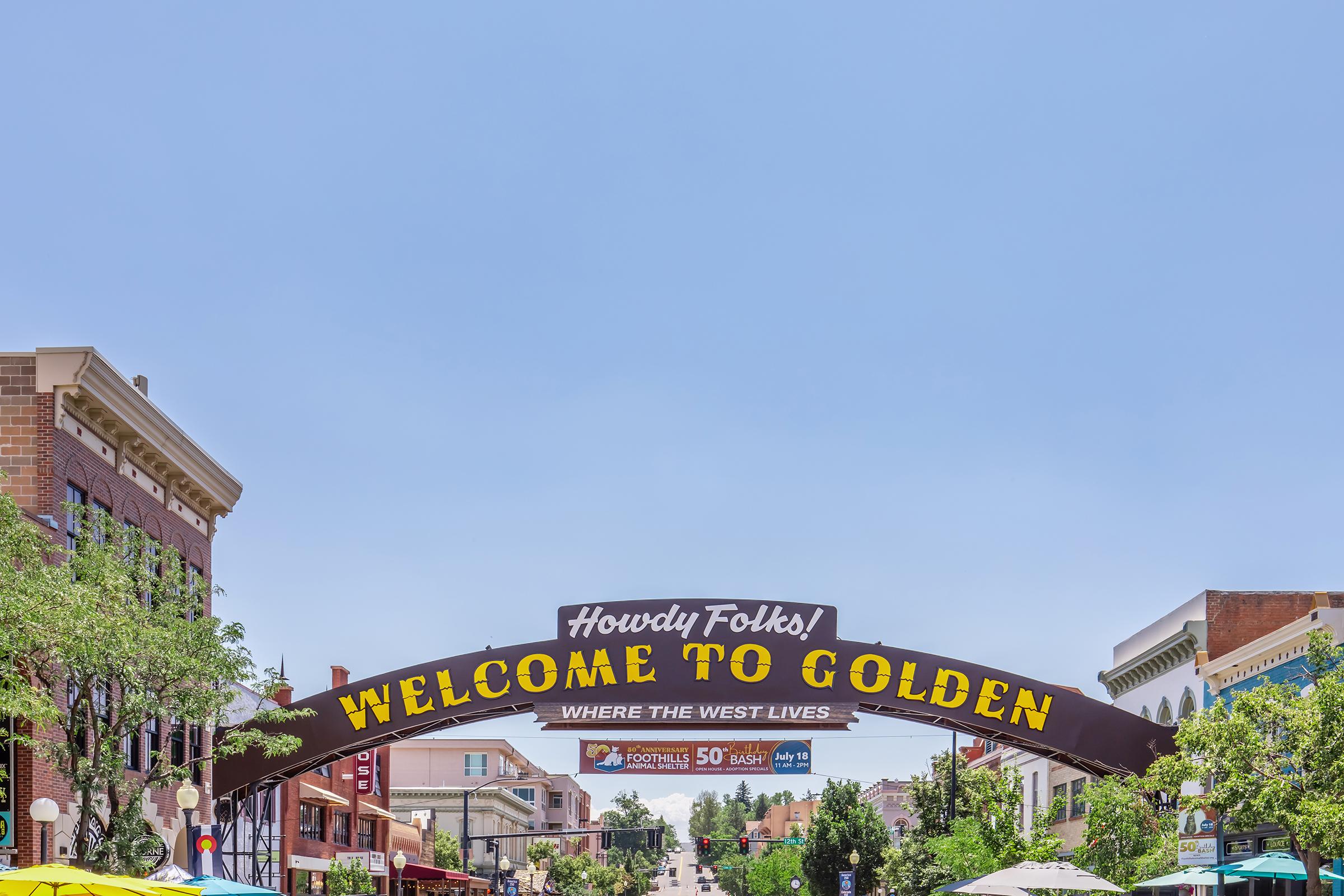 A large archway sign welcoming visitors to Golden, with the text "Howdy Folks! Welcome to Golden - Where the West Lives." The scene features buildings lining a street, with green trees and outdoor seating visible, under a clear blue sky.