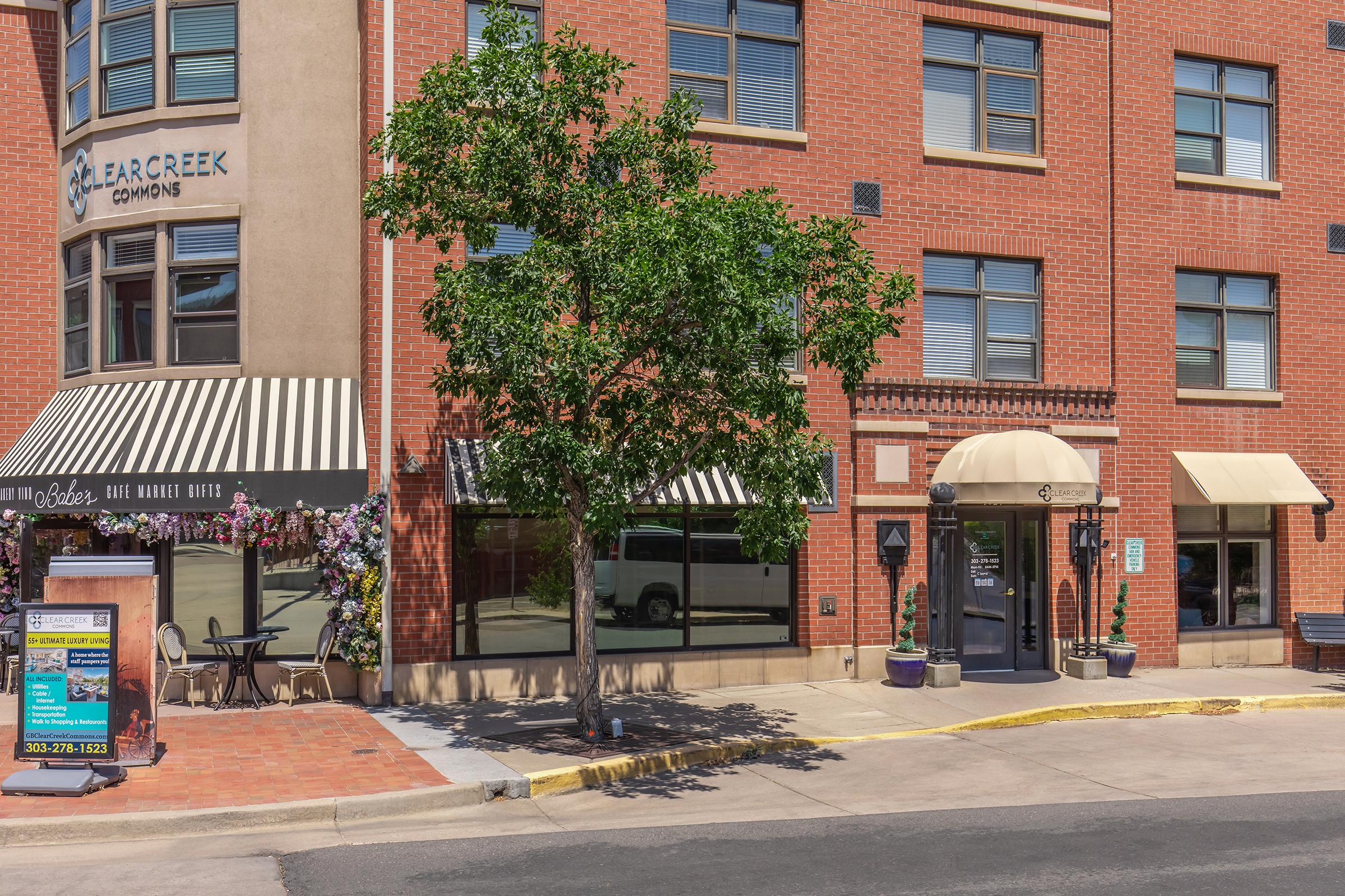 A view of a building named "Bear Creek Commons" featuring a storefront with striped awnings. There’s a landscaped area with a tree and some flowering plants. The pavement is made of bricks, and a sign for a nearby business is visible. The scene captures a sunny day in an urban setting.