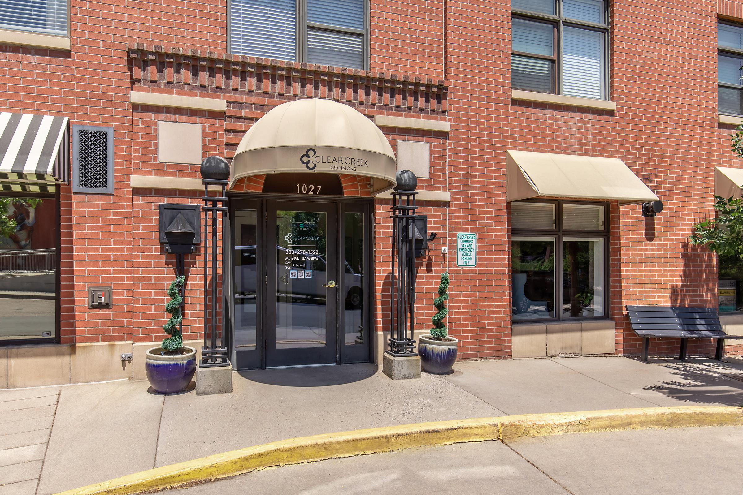 Entrance to a building with a "Clear Creek" sign above the door. The facade features red brick, a beige awning, decorative planters with greenery, and light fixtures on either side of the entrance. A bench is positioned nearby, and there are large windows visible on the ground floor.