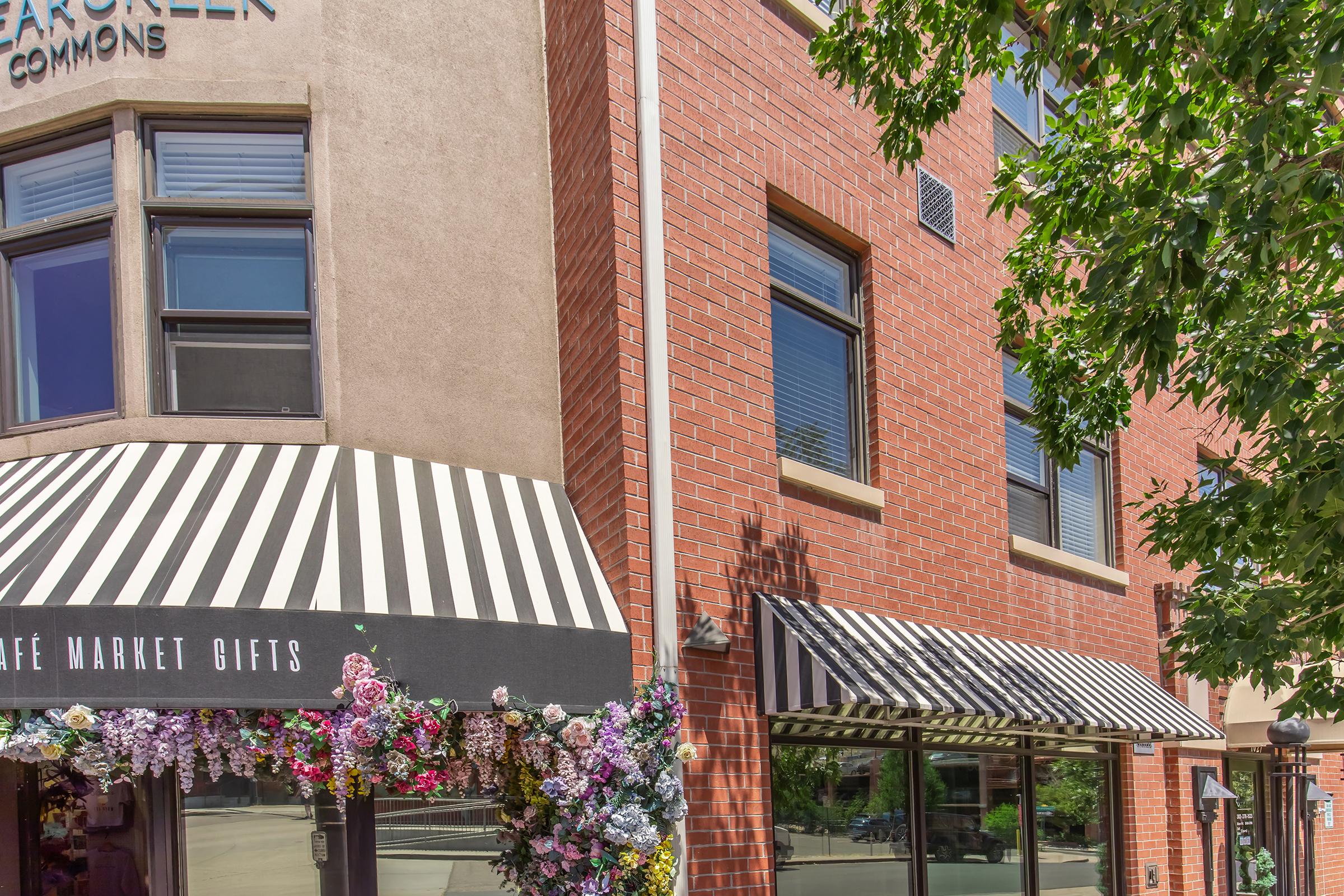 A brick building featuring large windows and striped awnings. One awning has the text "Café Market Gifts." The storefront is adorned with colorful floral decorations, adding a vibrant touch to the scene. Nearby trees provide shade, enhancing the inviting atmosphere of the urban setting.