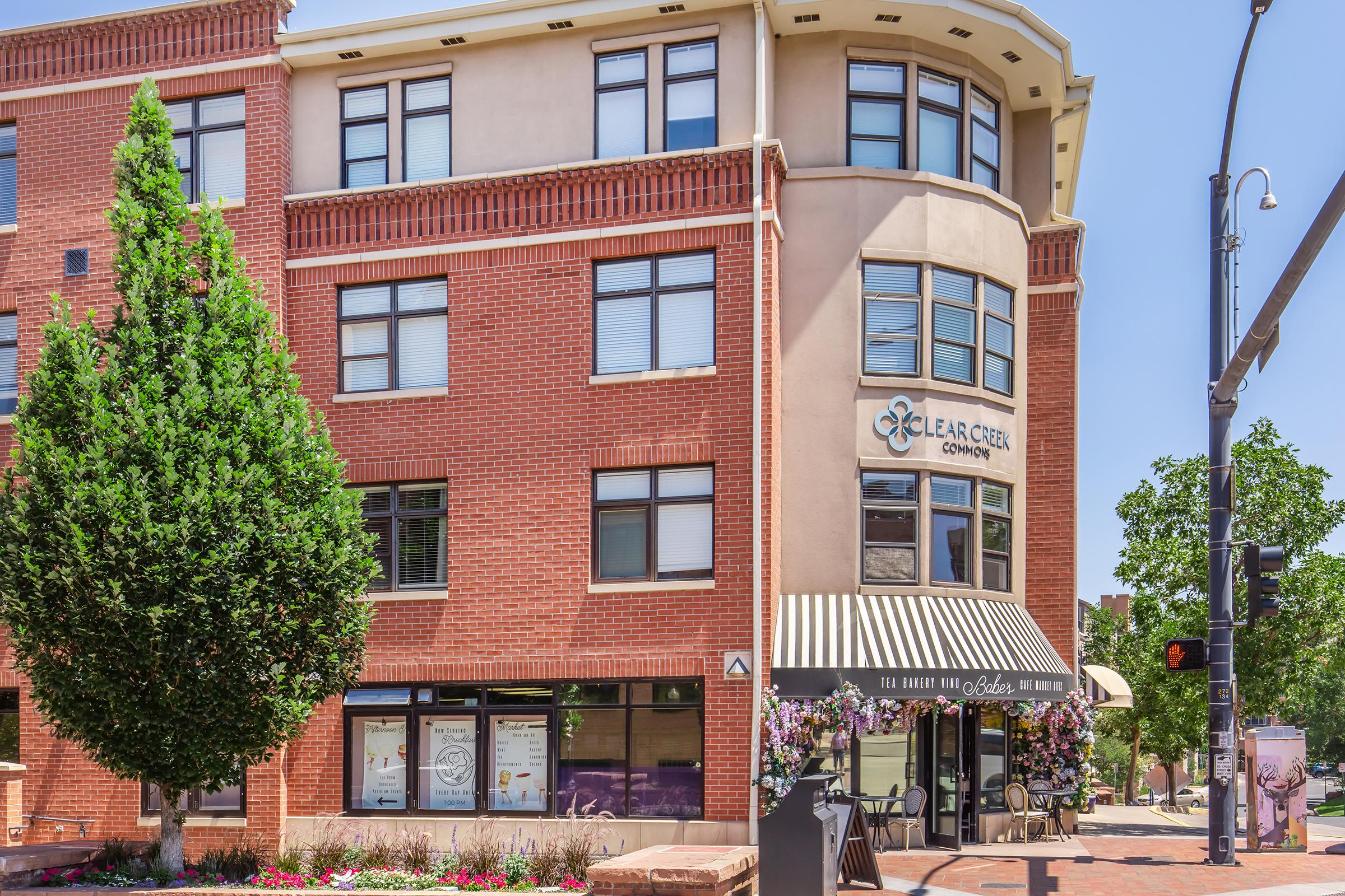 A brick building with large windows and a corner entrance, featuring a sign for "Clear Creek Commons." There are flowering plants in front, a tree beside the building, and street signs visible. The scene is set under a clear blue sky, suggesting a sunny day.