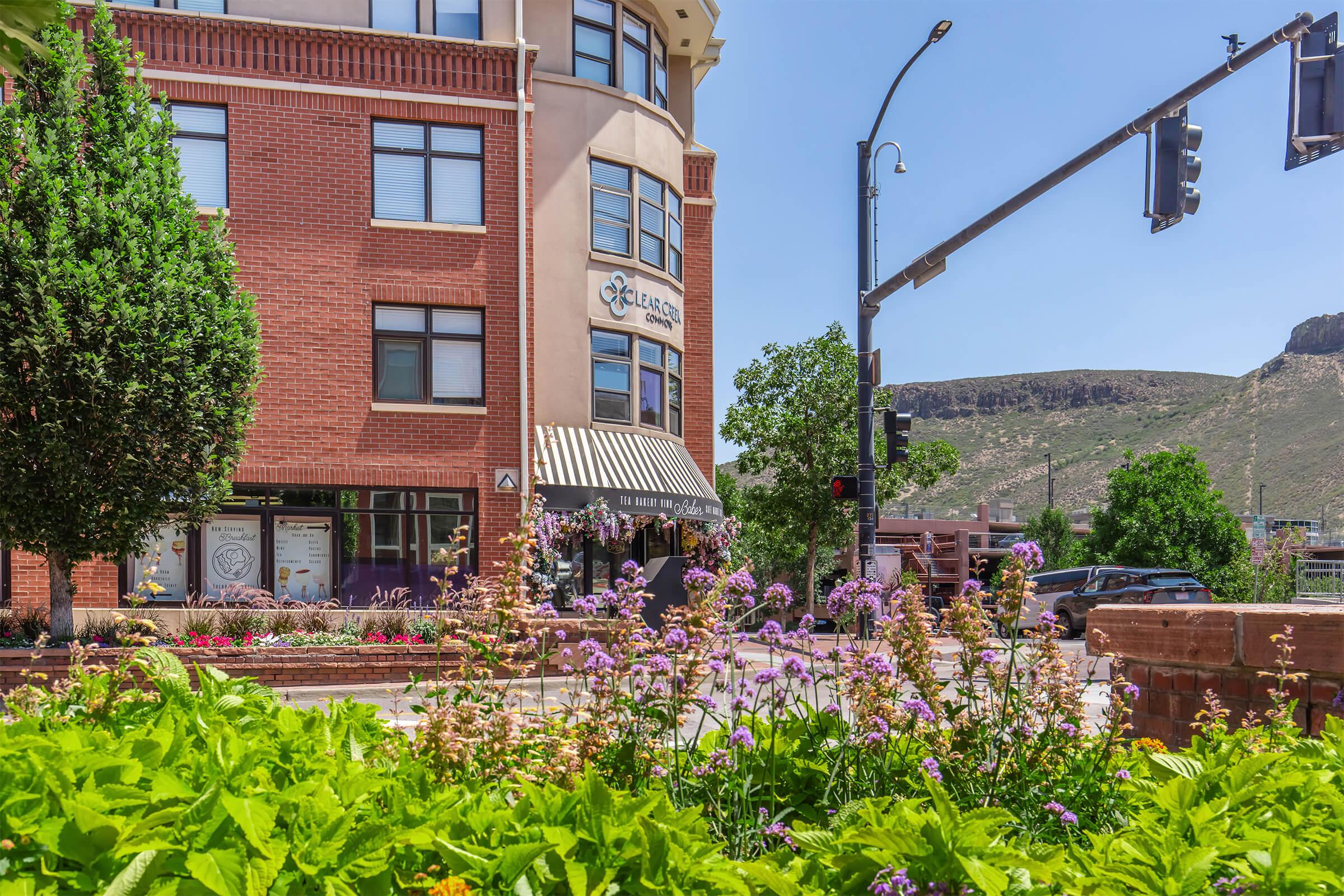 A scenic view of a street corner featuring a brick building with a striped awning labeled "Klein's" and a floral landscape in the foreground. Green trees surround the area, and a traffic light is visible, with rocky hills in the background under a clear blue sky.