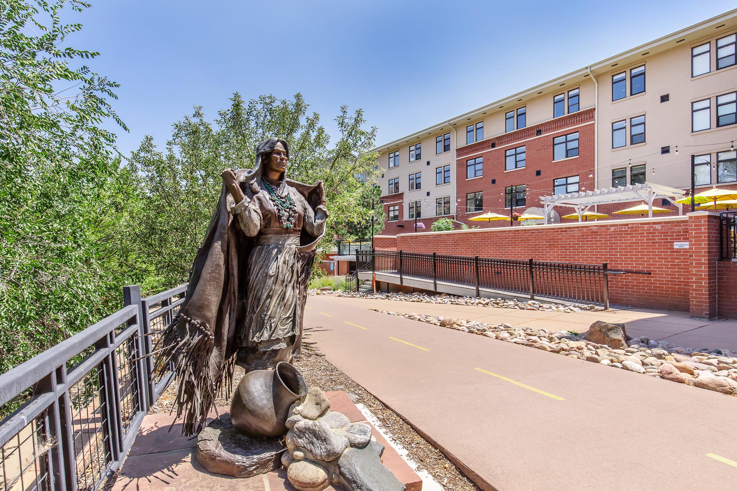 Statue of a Native American woman wearing traditional attire, holding a pot and standing on a pathway lined with rocks. In the background, there is a modern multi-story building with large windows and outdoor seating. Lush greenery surrounds the area, creating a serene atmosphere.