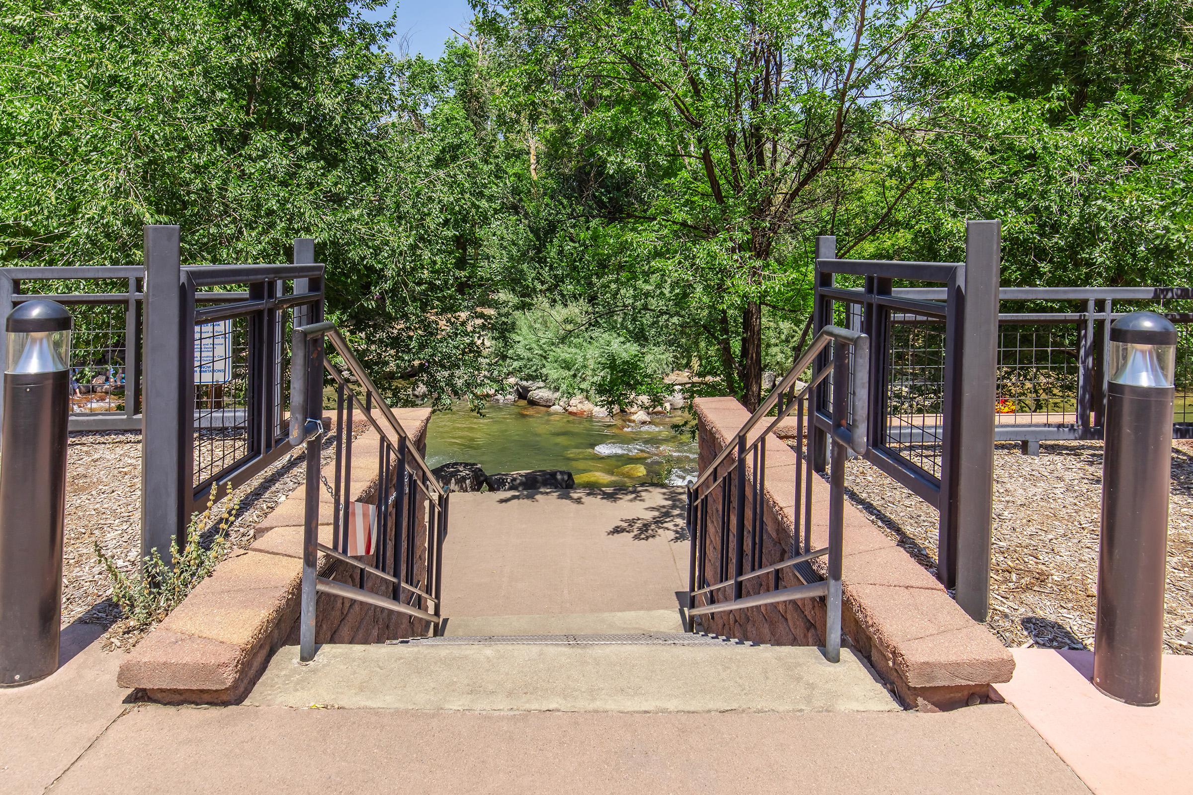 A pathway leading down a set of stairs towards a small stream, surrounded by green trees and shrubs. The area features metal railings and light posts on either side of the stairs, providing a serene natural setting for visitors.
