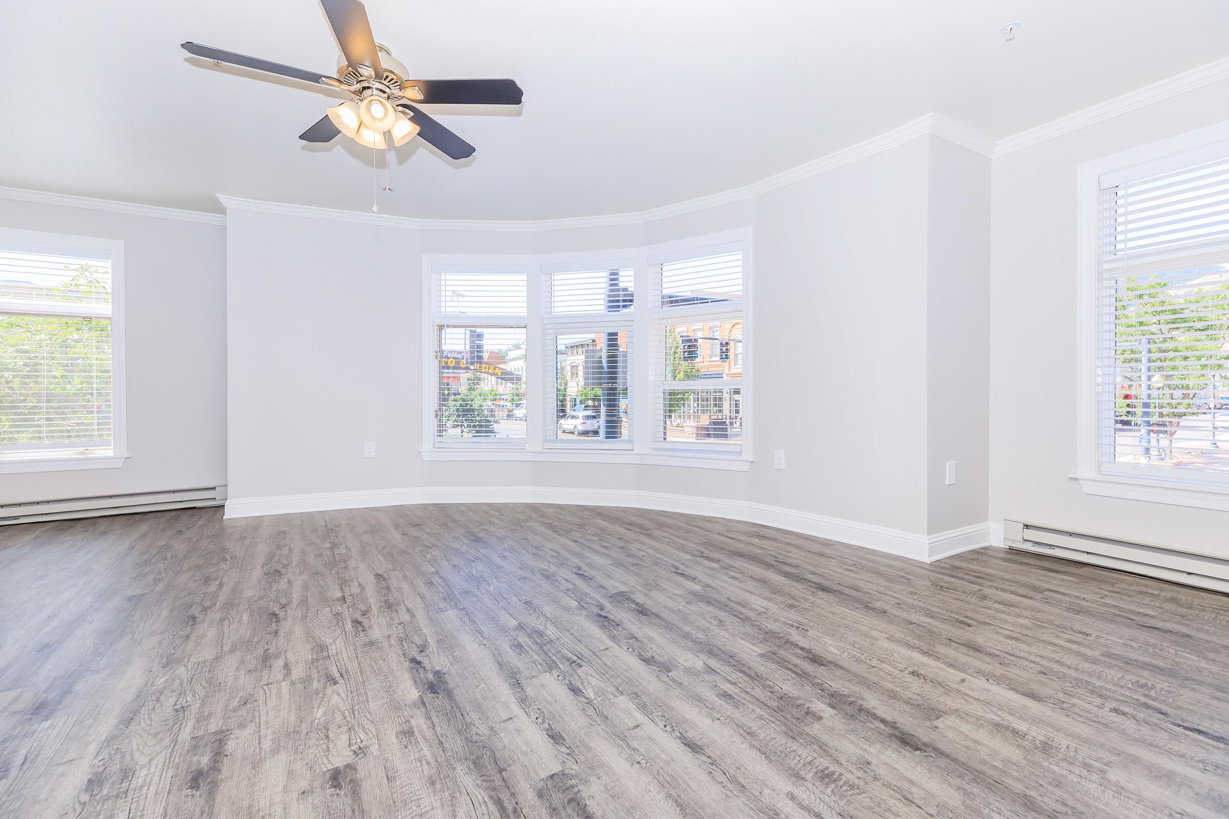 A spacious, empty living room with light-colored walls and hardwood flooring. Large bay windows let in natural light, and a ceiling fan is installed. The room has a modern and airy feel, making it suitable for various furniture arrangements.