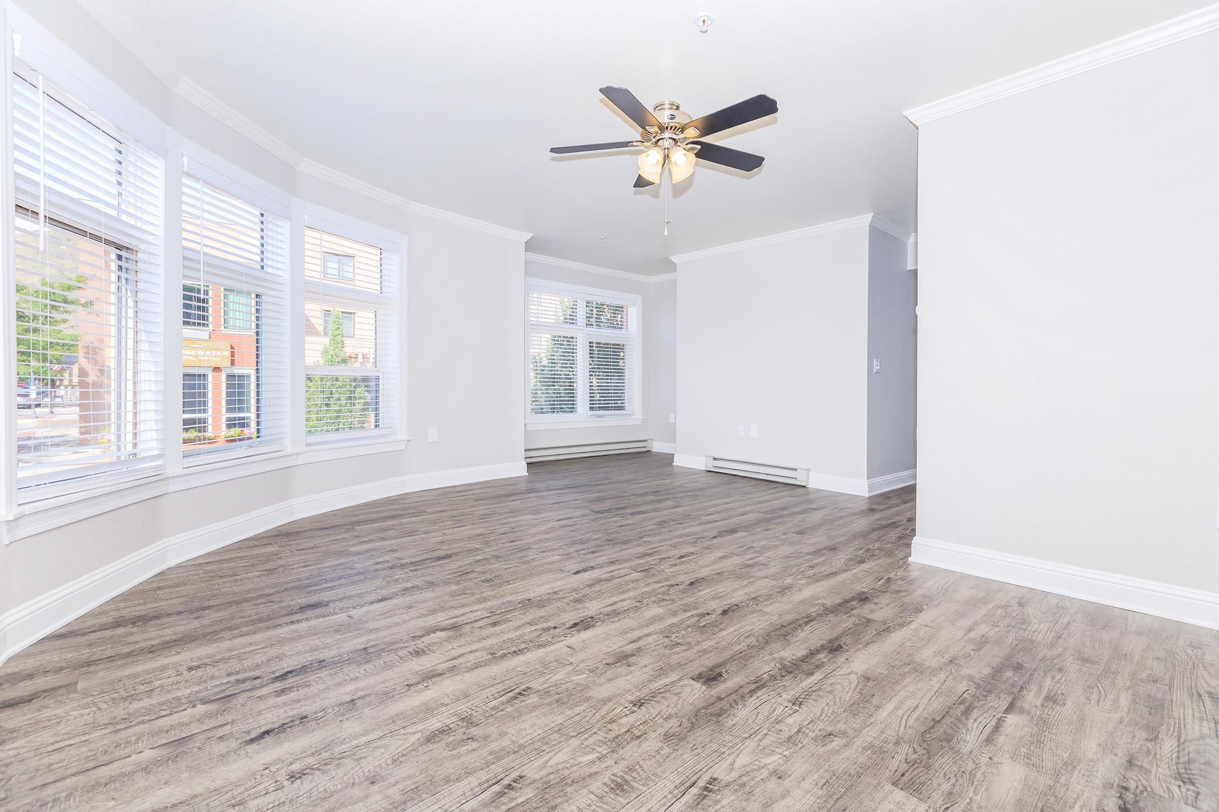 A spacious, empty living room featuring light-colored walls, large windows with white blinds, and a wooden floor. A ceiling fan with light fixtures hangs from the ceiling. The room has a bright and airy feel, with natural light streaming in from the windows.