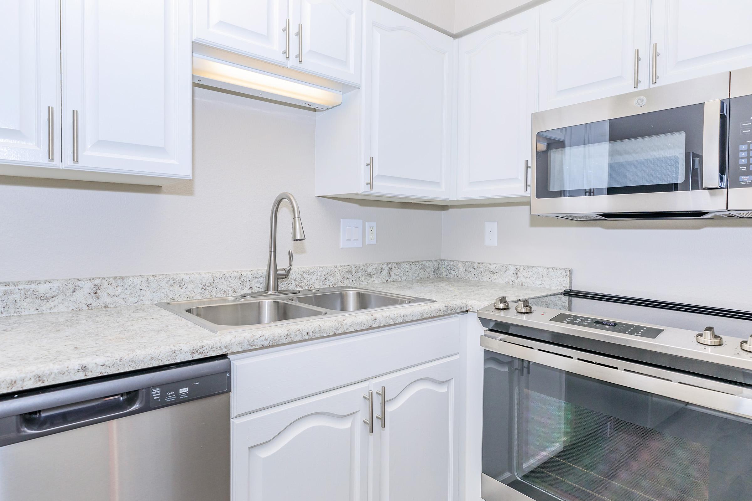 Modern kitchen with white cabinetry, granite countertops, a double sink, a stainless steel dishwasher, and an oven with a microwave above. The layout is clean and contemporary, featuring bright lighting and minimalistic design elements.