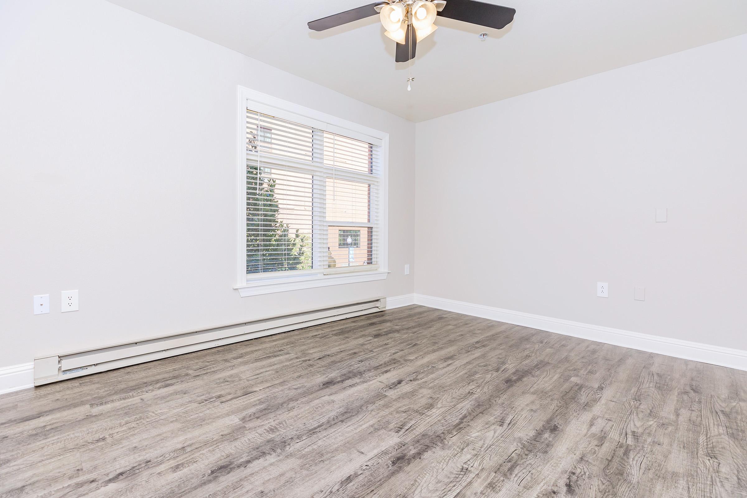 Empty room with light gray walls and a ceiling fan, featuring a large window with white blinds allowing natural light. The floor is covered with light wood-like laminate. There are baseboard heaters beneath the window and minimal decor, creating a bright and open feel.