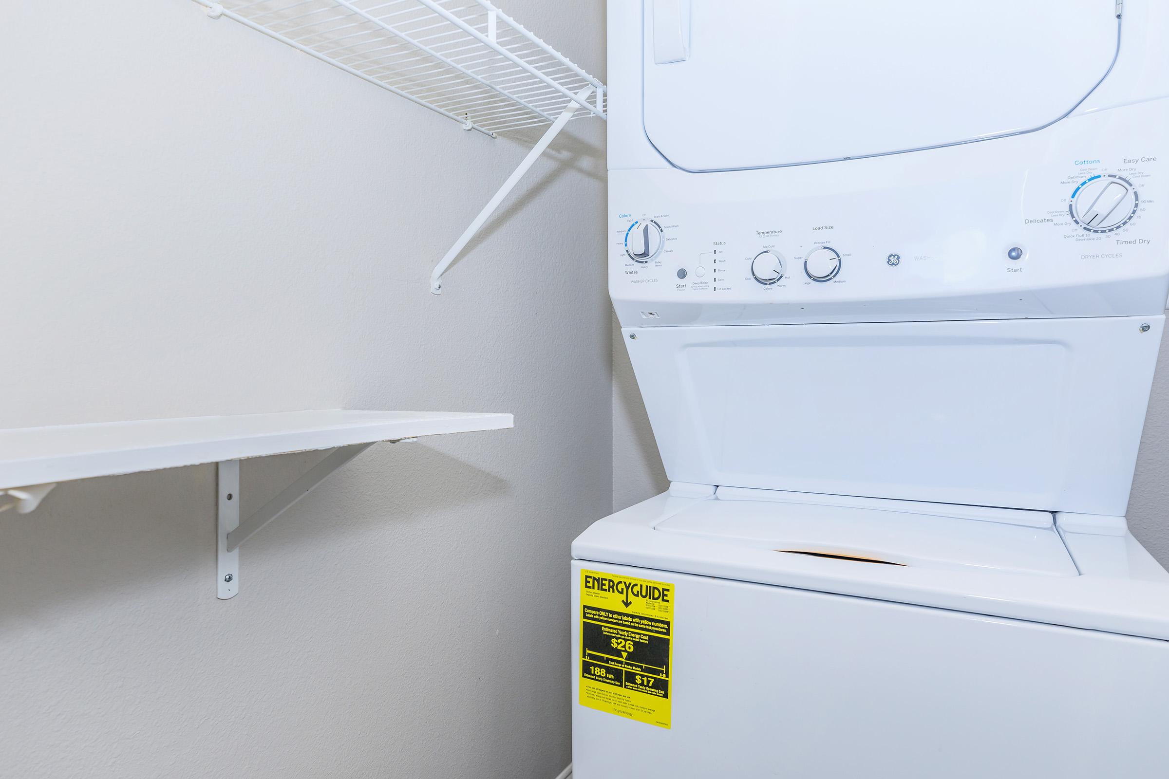 A white stacked washer and dryer unit is placed against a gray wall. A shelf is mounted above the washer, providing additional storage space. The energy guide label is visible on the dryer, indicating its energy efficiency.