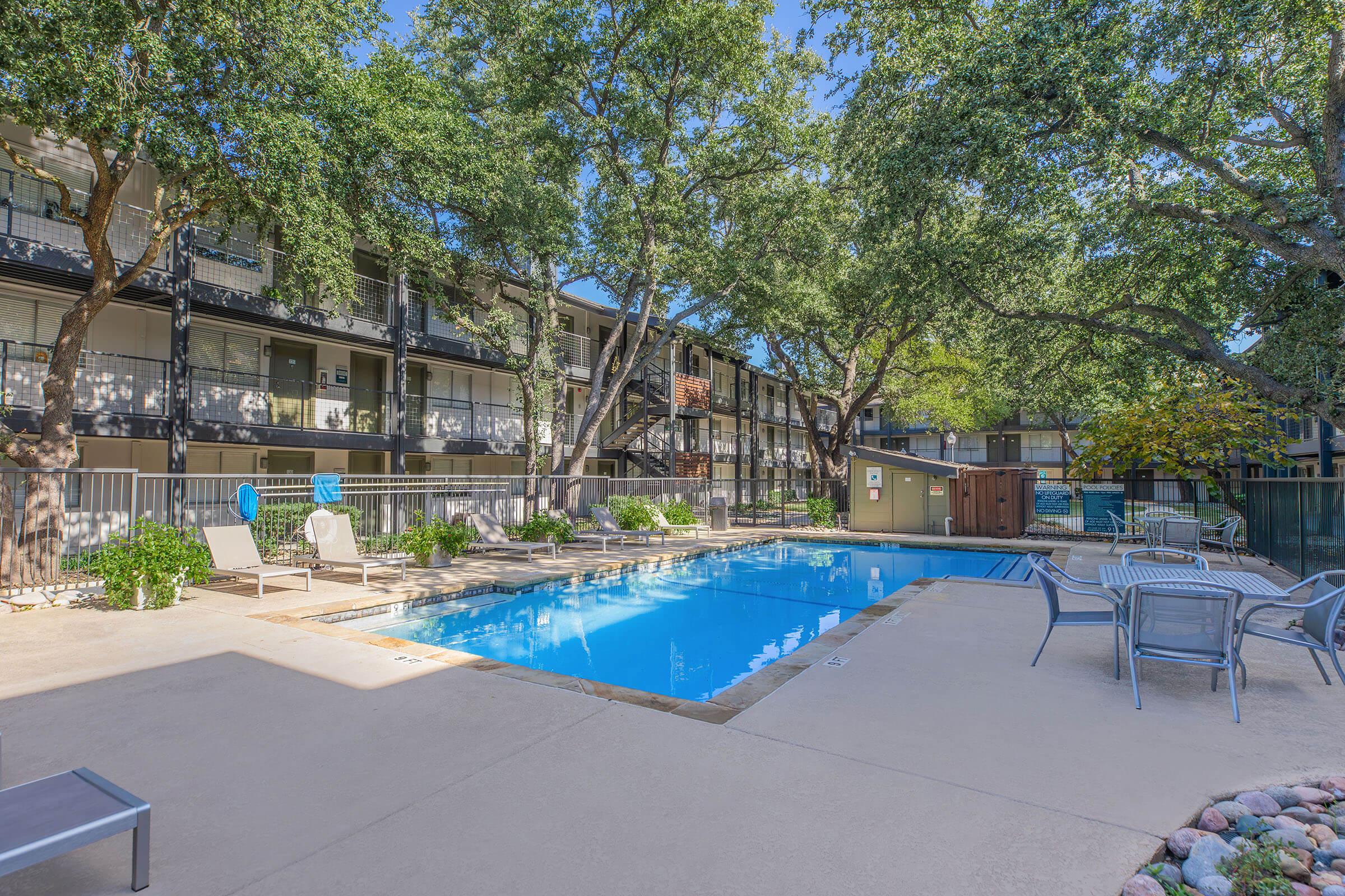 A residential area featuring a swimming pool surrounded by lounge chairs, green landscaping, and several trees. In the background, there are multi-story apartment buildings with balconies. The scene is bright and well-maintained, suggesting a relaxing outdoor space for residents.