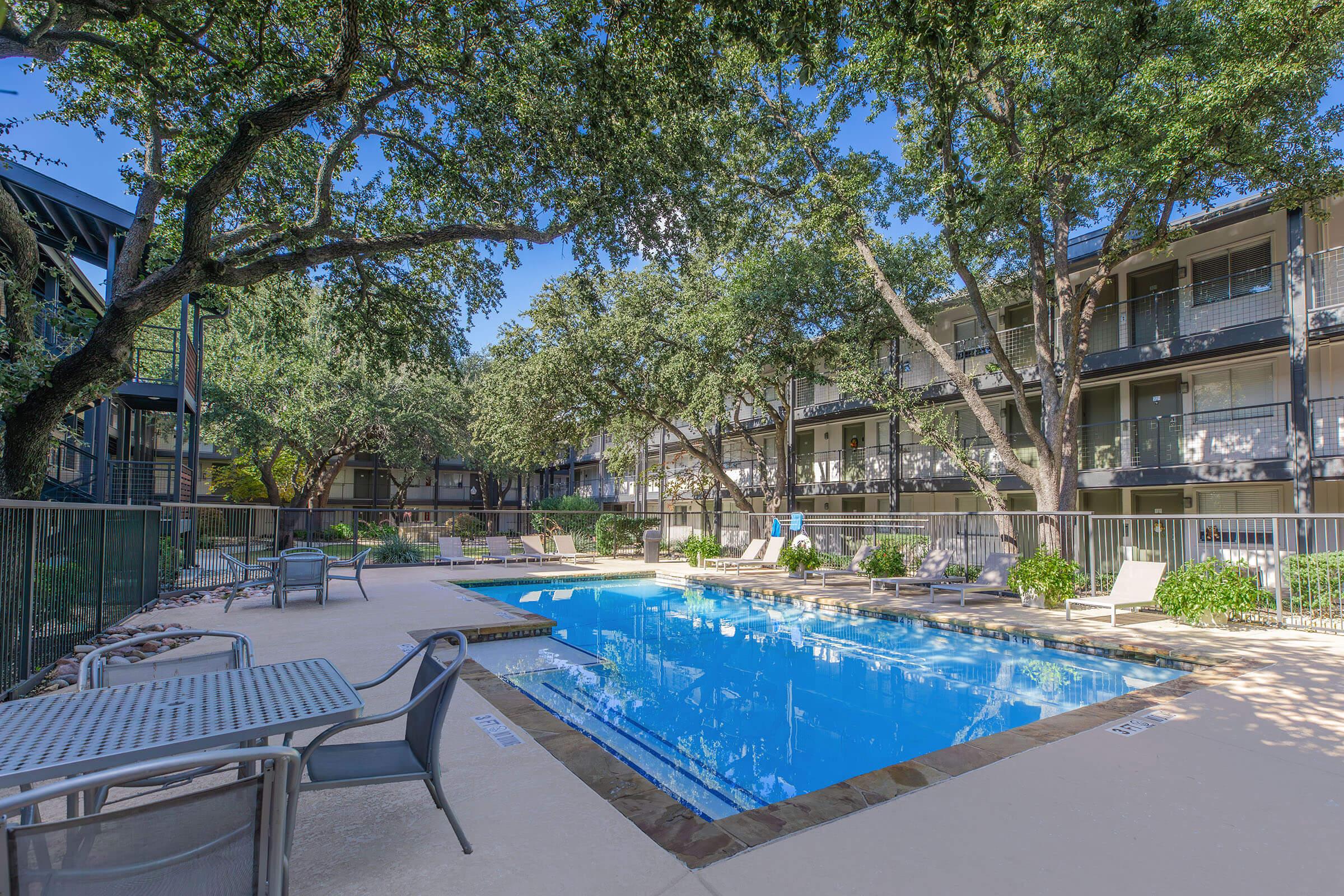 A sunny pool area surrounded by trees, featuring a clear blue pool with lounge chairs positioned around it. There are tables with chairs in the foreground, and multi-story apartment buildings with balconies in the background. The setting is relaxing and inviting, ideal for leisure.