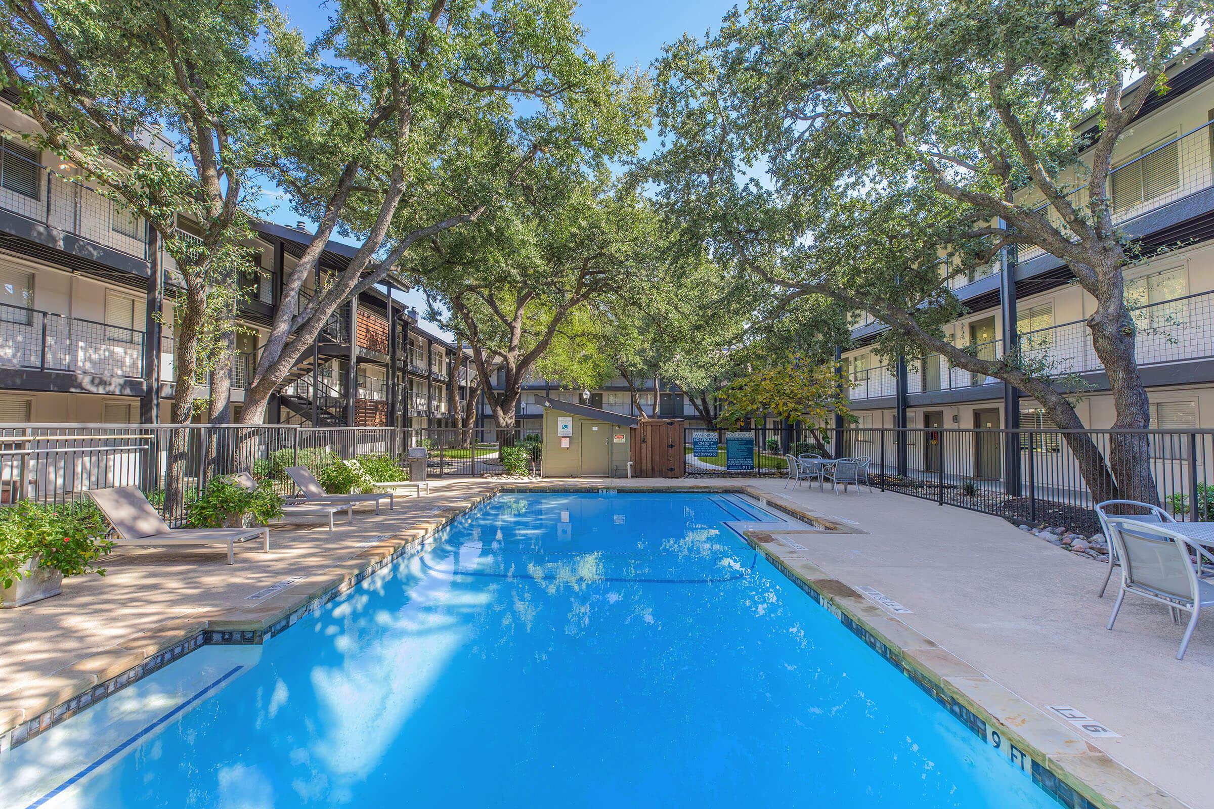A clear swimming pool surrounded by trees and greenery, with multiple lounge chairs and a light-colored patio area. There are two-story apartment buildings in the background, with balconies visible. The scene is bright and inviting, suggesting a relaxing outdoor atmosphere.