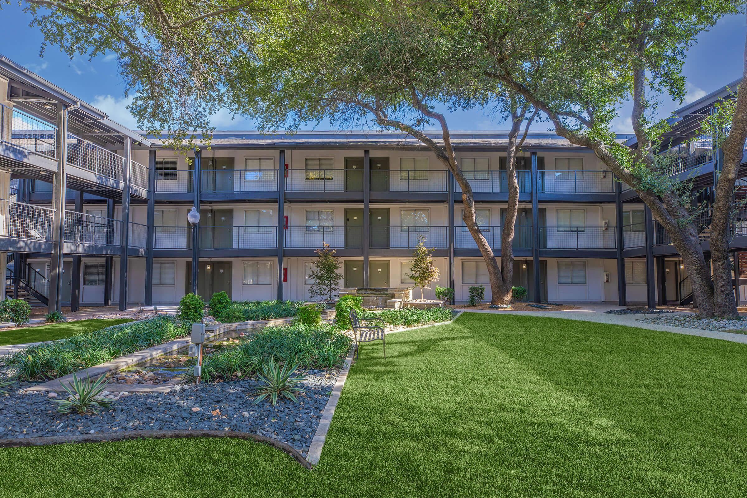 A well-maintained outdoor courtyard of an apartment complex featuring a green lawn, neatly arranged shrubs, a stone pathway, and several trees. The buildings surround the courtyard with multiple levels, showcasing balconies and patios along the exterior. Blue skies are visible above.