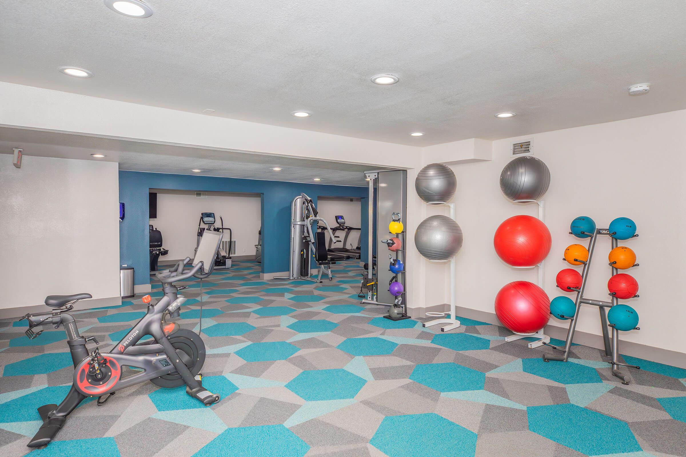 A modern gym space featuring exercise equipment. The room has a stationary bike, various exercise balls stacked, and free weights. The floor is designed with a geometric pattern in shades of gray and teal. In the background, additional workout machines are visible, suggesting a well-equipped fitness area.