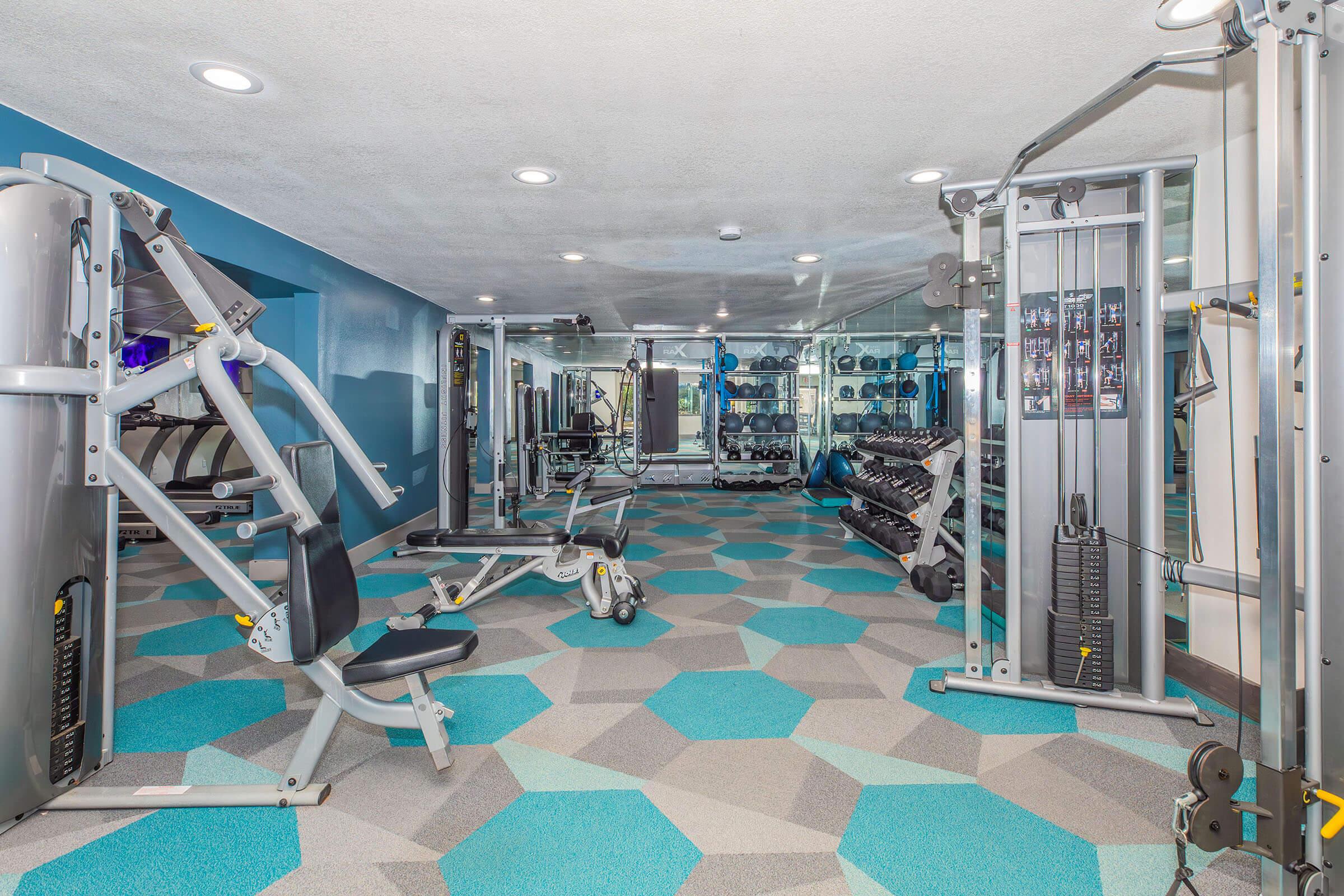 A modern gym interior featuring various exercise equipment, including weight machines, free weights, and cardio machines. The space has a blue and gray color scheme with geometric-patterned flooring, bright lighting, and mirrors reflecting the workout areas.