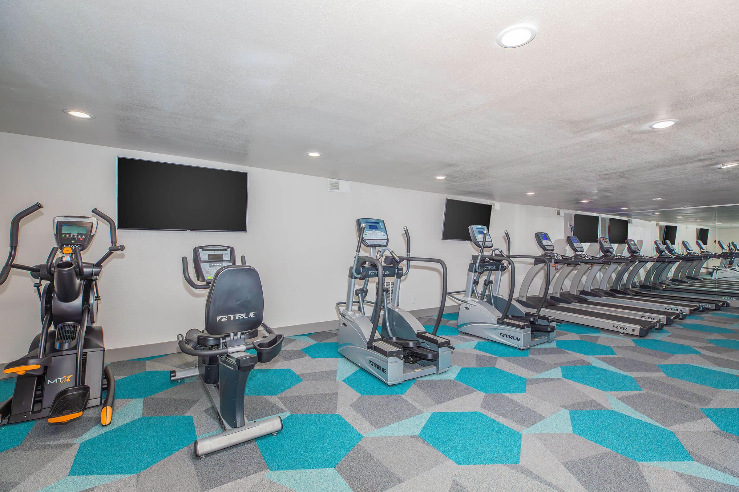 A modern fitness room featuring several exercise machines including ellipticals, treadmills, and a stationary bike. The room has a sleek design with bright blue and gray geometric flooring, large mirrors, and flat-screen TVs mounted on the walls. The space is well-lit and inviting for workouts.