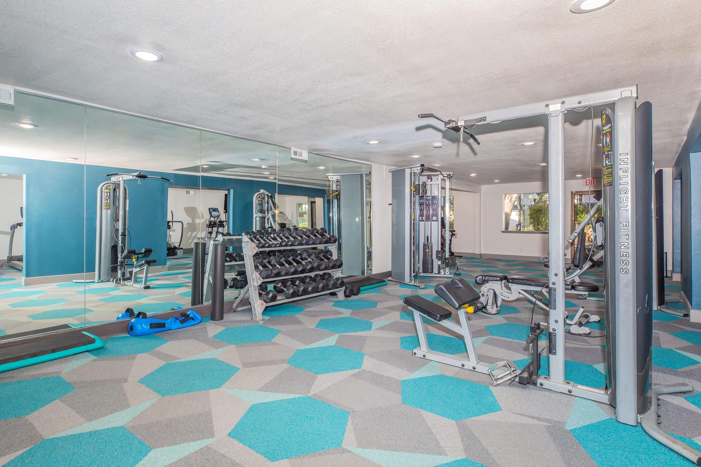 A modern gym interior featuring various fitness equipment, including weight machines, free weights, and cardio machines. The space is brightly lit, with mirrors on one wall and a colorful hexagonal pattern on the floor.