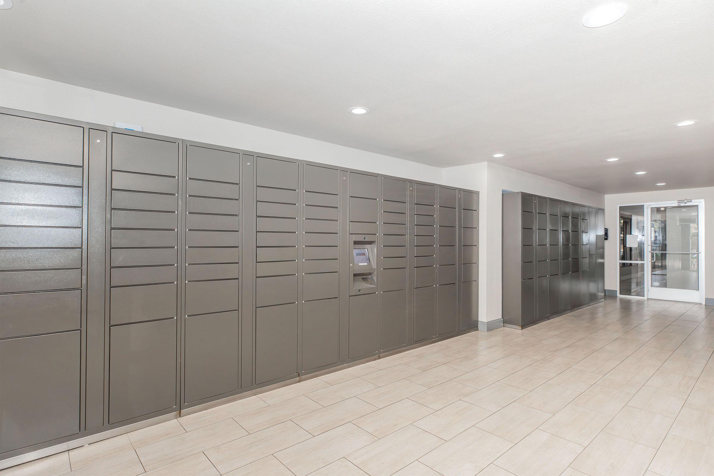 A corridor featuring a series of dark gray lockers with horizontal slats, designed for package delivery. The floor is tiled in light colors, and there is a visible entrance to the right, providing access to another area. Soft overhead lighting enhances the modern and clean aesthetic.