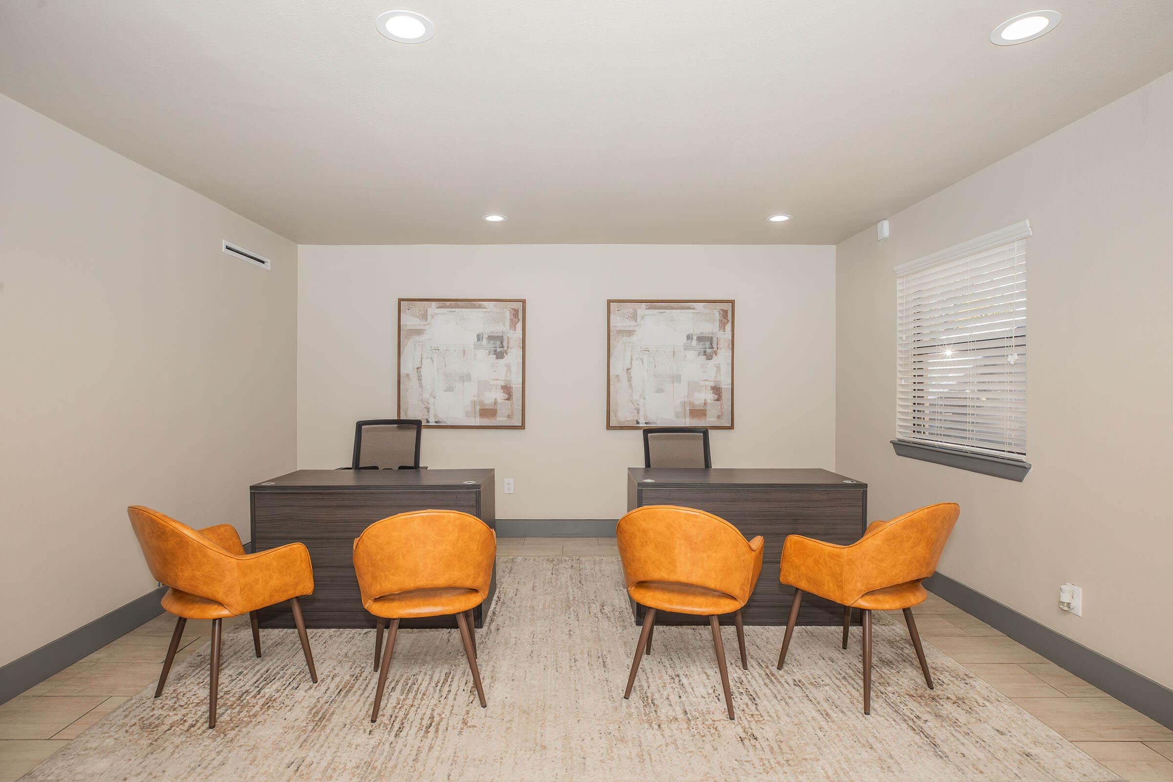 A modern office space featuring two desks with black chairs and four orange upholstered chairs arranged in front. Light-colored walls and a neutral rug complement the decor. Two abstract paintings are mounted on the wall, adding a touch of artistry to the professional environment. Bright, overhead lighting illuminates the area.