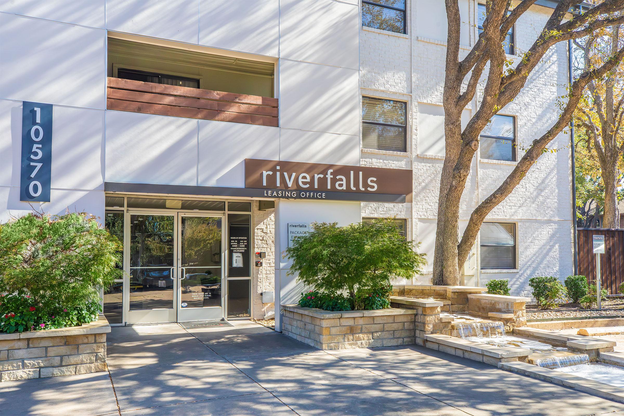 Leasing office entrance for Riverfalls, featuring a modern design with a sign that reads "riverfalls Leasing Office." The entrance is surrounded by trees and landscaping, with stone pathway and structure details. The building exterior is light-colored and contemporary.