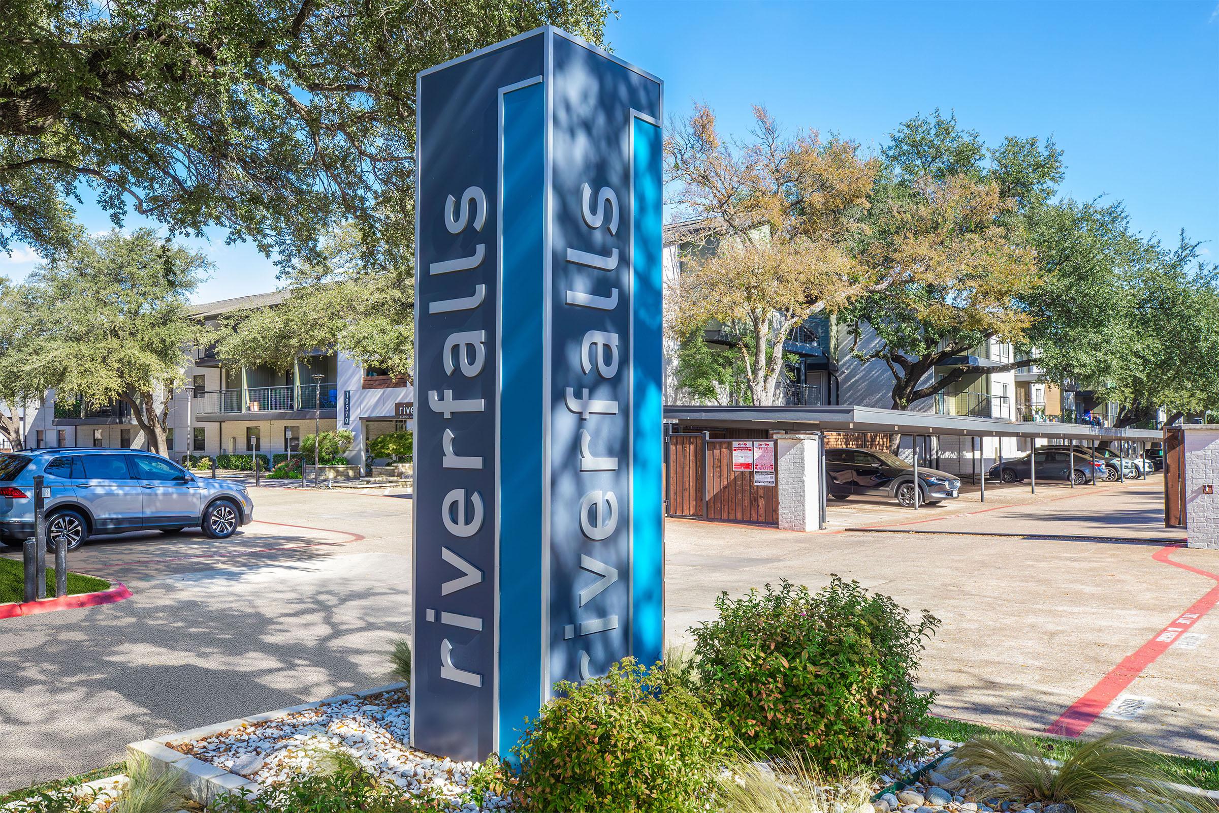 Sign for Riverfalls apartments surrounded by lush greenery and trees, with a parking area and several parked cars visible. The building in the background showcases modern architecture against a clear blue sky.