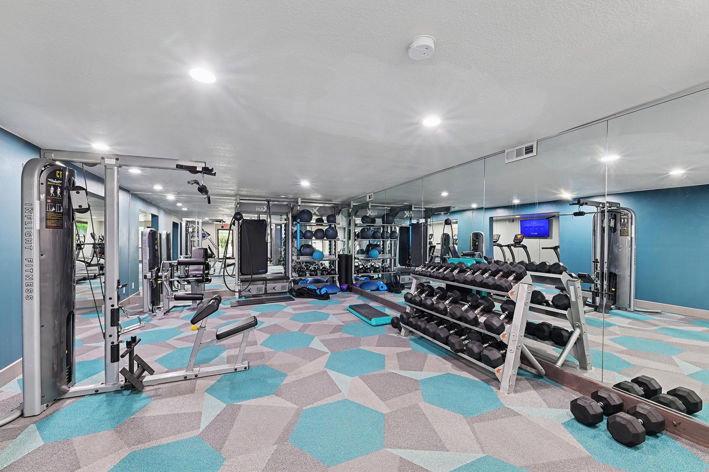 A modern gym interior featuring multifunctional workout equipment, including weight machines and free weights. The walls are mirrored, and vibrant teal and gray flooring adds a contemporary touch. There's ample space for exercise with a flat-screen TV on the wall.