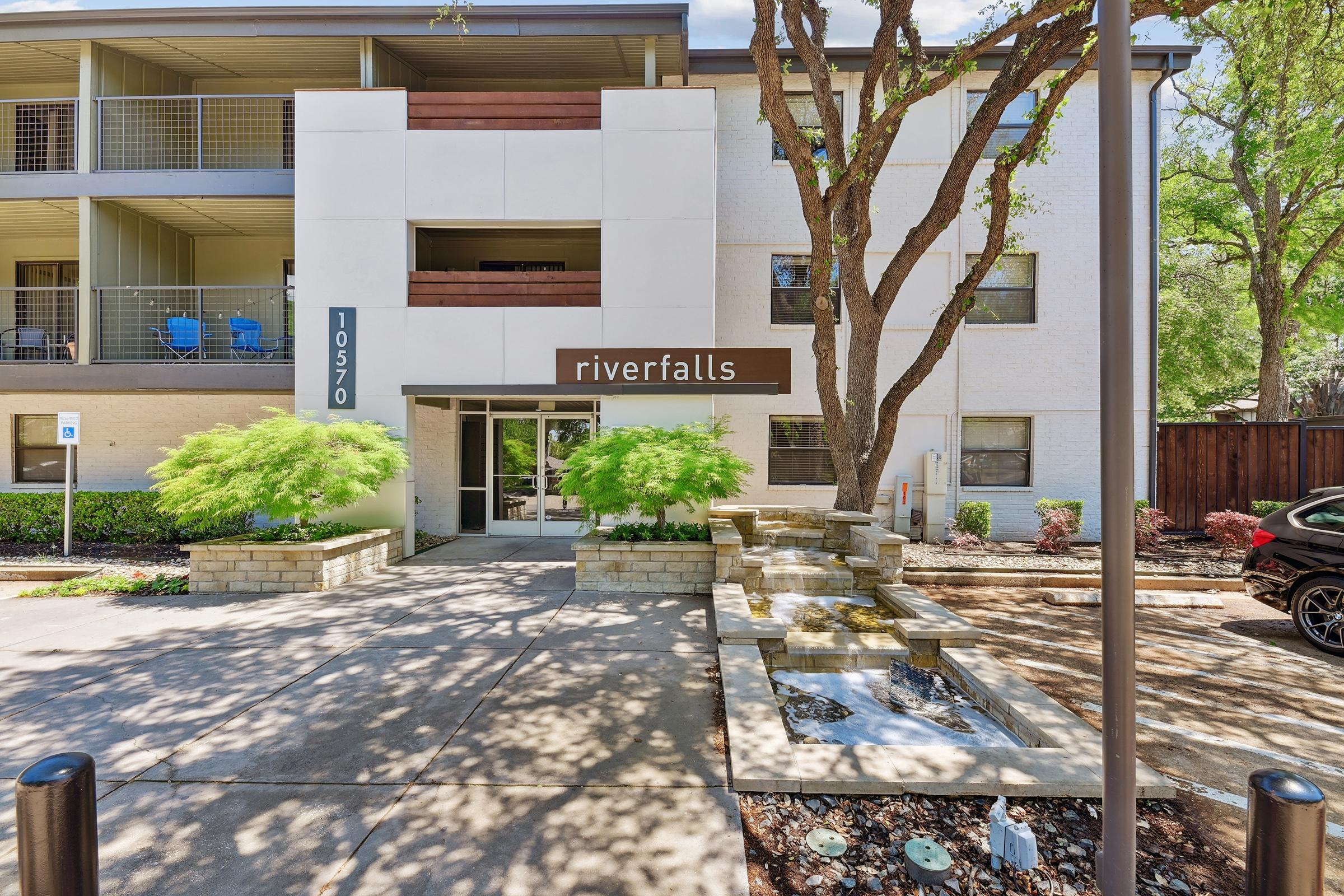 Exterior view of the Riverfalls apartment building with a modern design featuring a white facade and brown accents. A small water fountain and landscaped greenery are in front, providing an inviting entrance. There are parked cars and a walkway leading to the entrance.