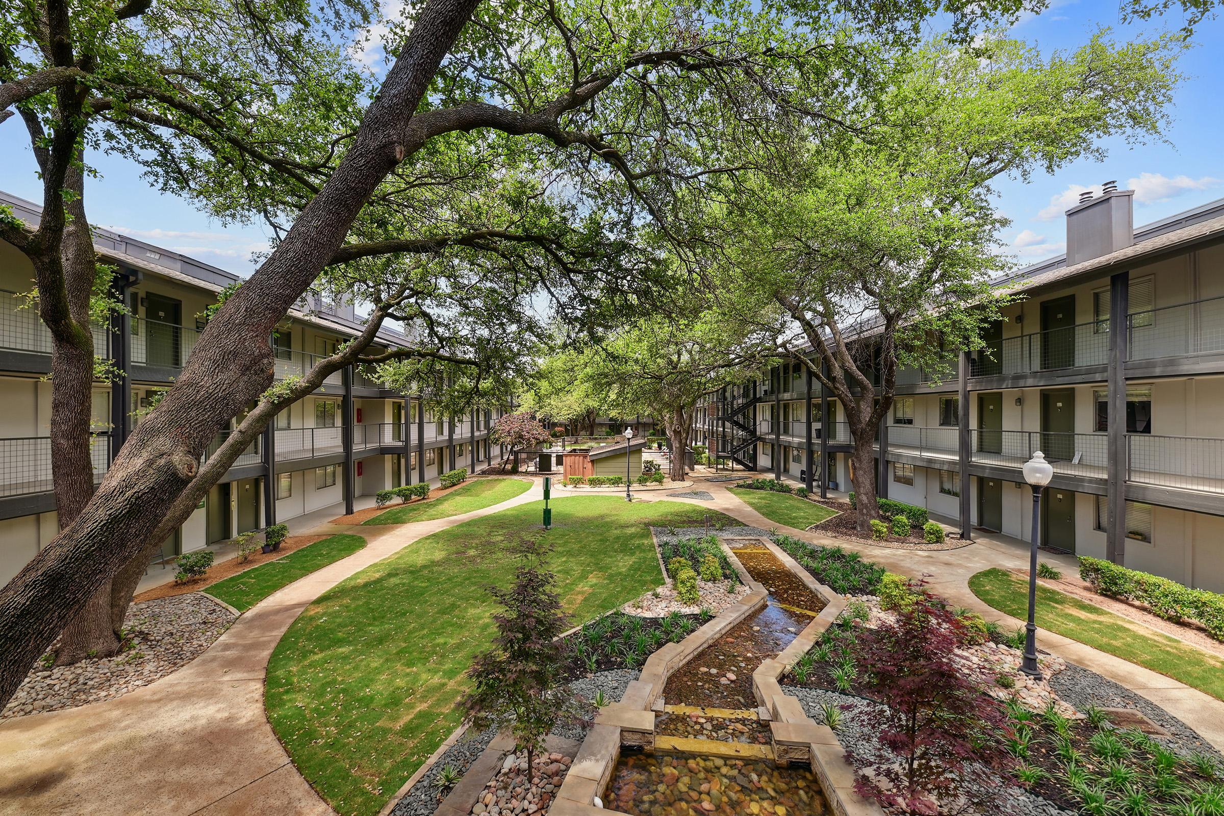 A landscaped courtyard surrounded by two-story apartment buildings. The area features a small water feature with a stream and rocks, lush green grass, and various shrubs and trees providing shade. The sky is clear with a few clouds, creating a tranquil and inviting atmosphere.
