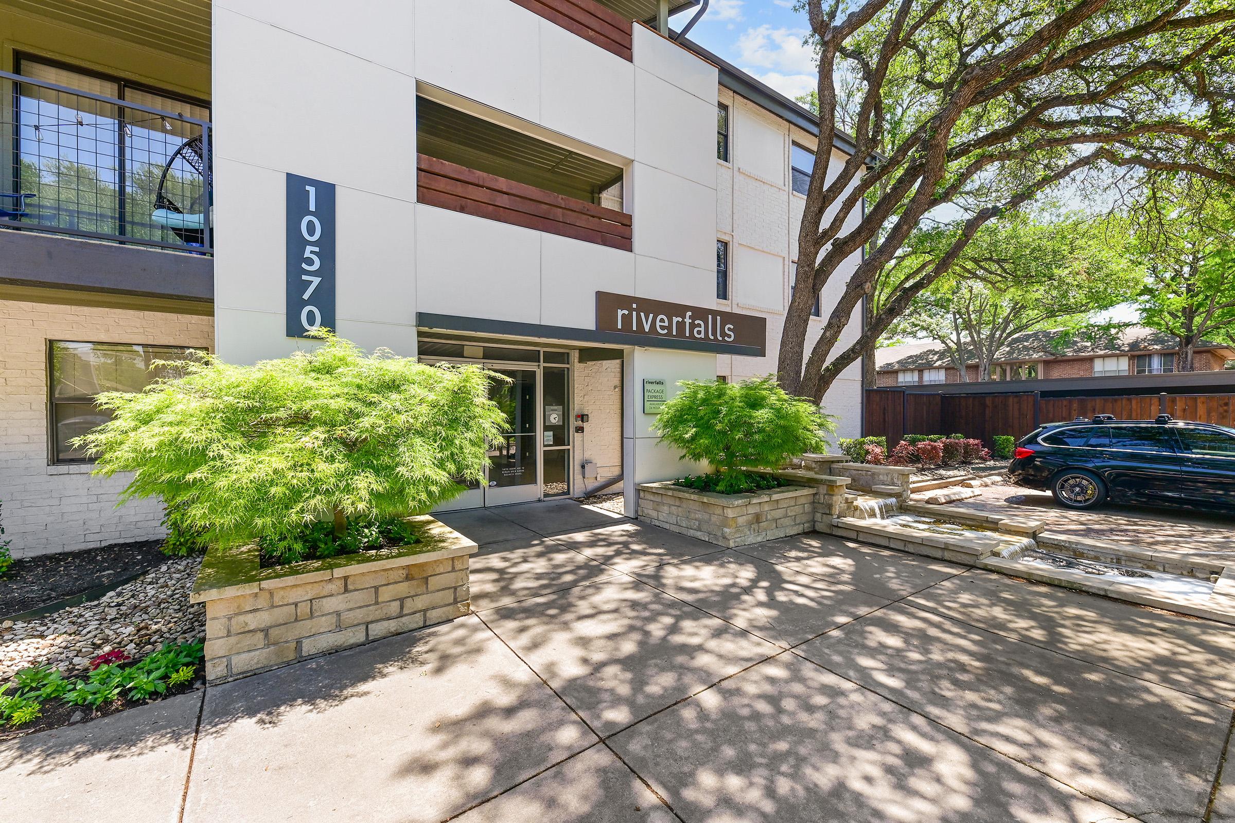 Entrance to an apartment building named "riverfalls," featuring a modern facade with large windows. The area is landscaped with green shrubs and flowers, and there’s a parked car nearby. The building has a welcoming atmosphere with trees providing shade.
